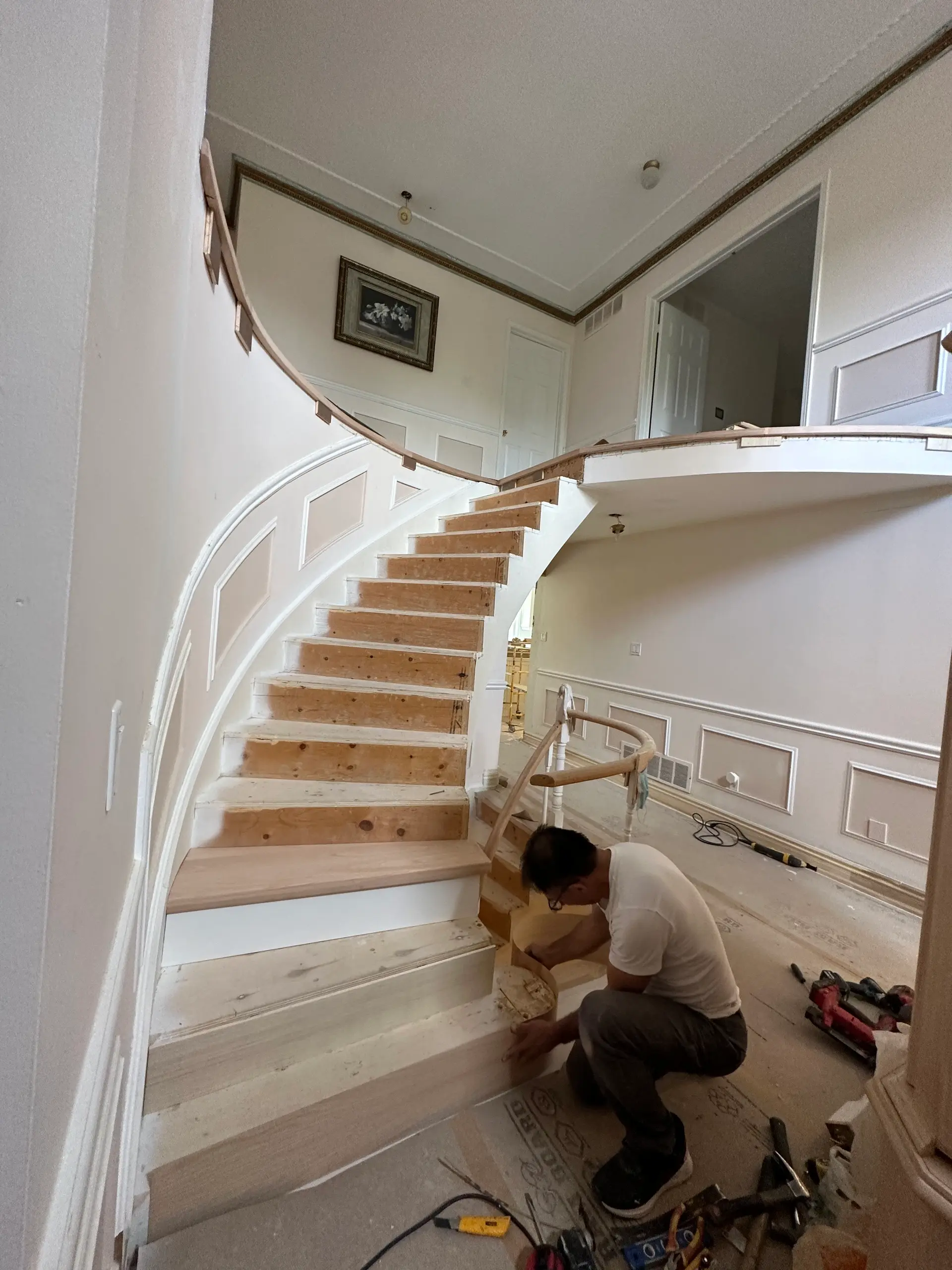 Curved wooden staircase under renovation, with a craftsman working on the finishing touches in a classic interior with wall moldings.