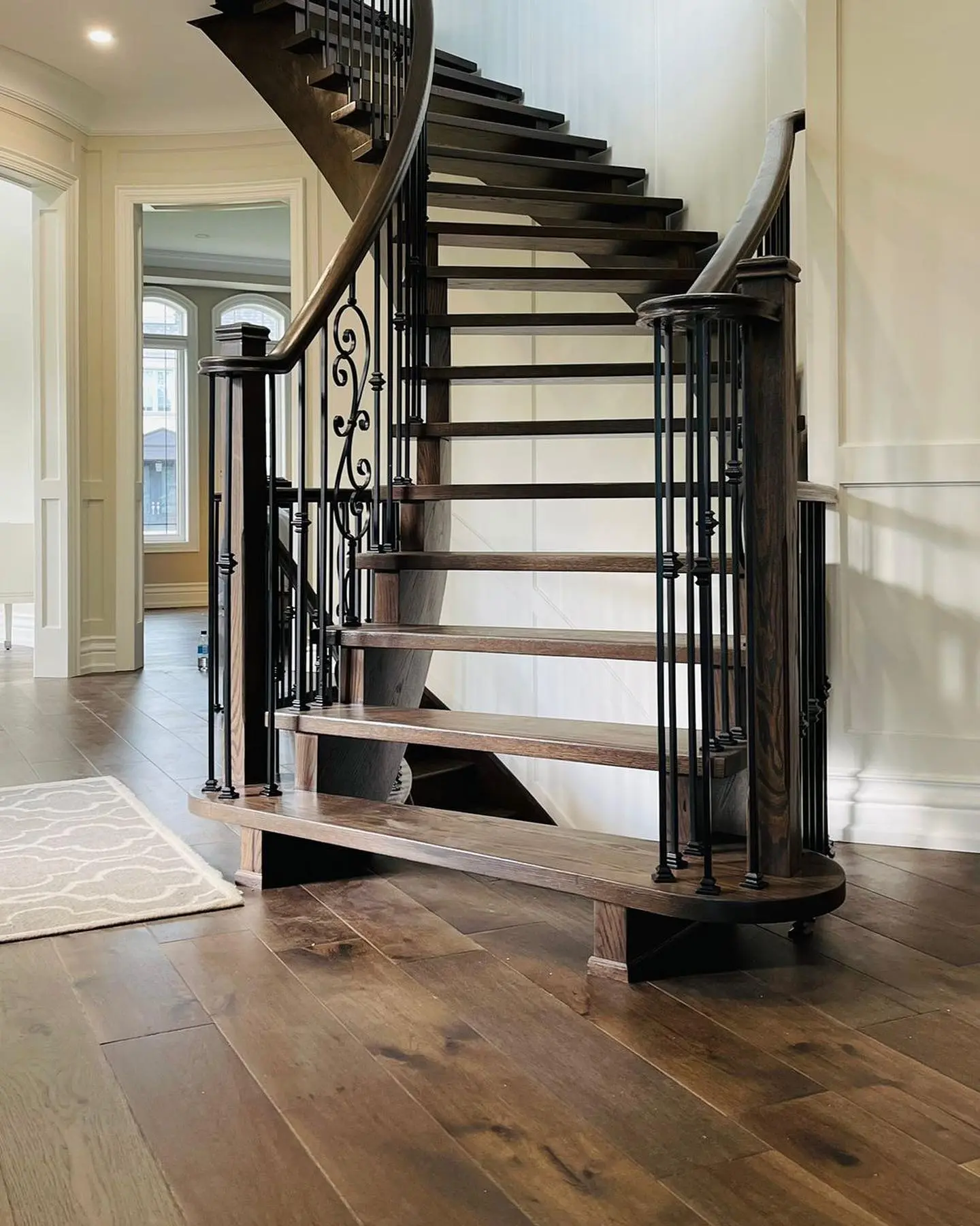 Escalier en bois courbé avec garde-corps en fer forgé, installé dans un intérieur élégant avec un sol en bois massif et des moulures murales.