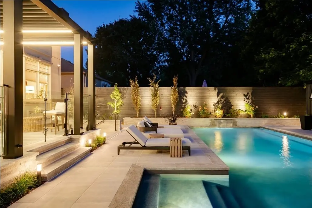 Modern backyard at dusk with illuminated pool, slab terrace, lounge chairs, wood fence, landscaping, and outdoor lighting