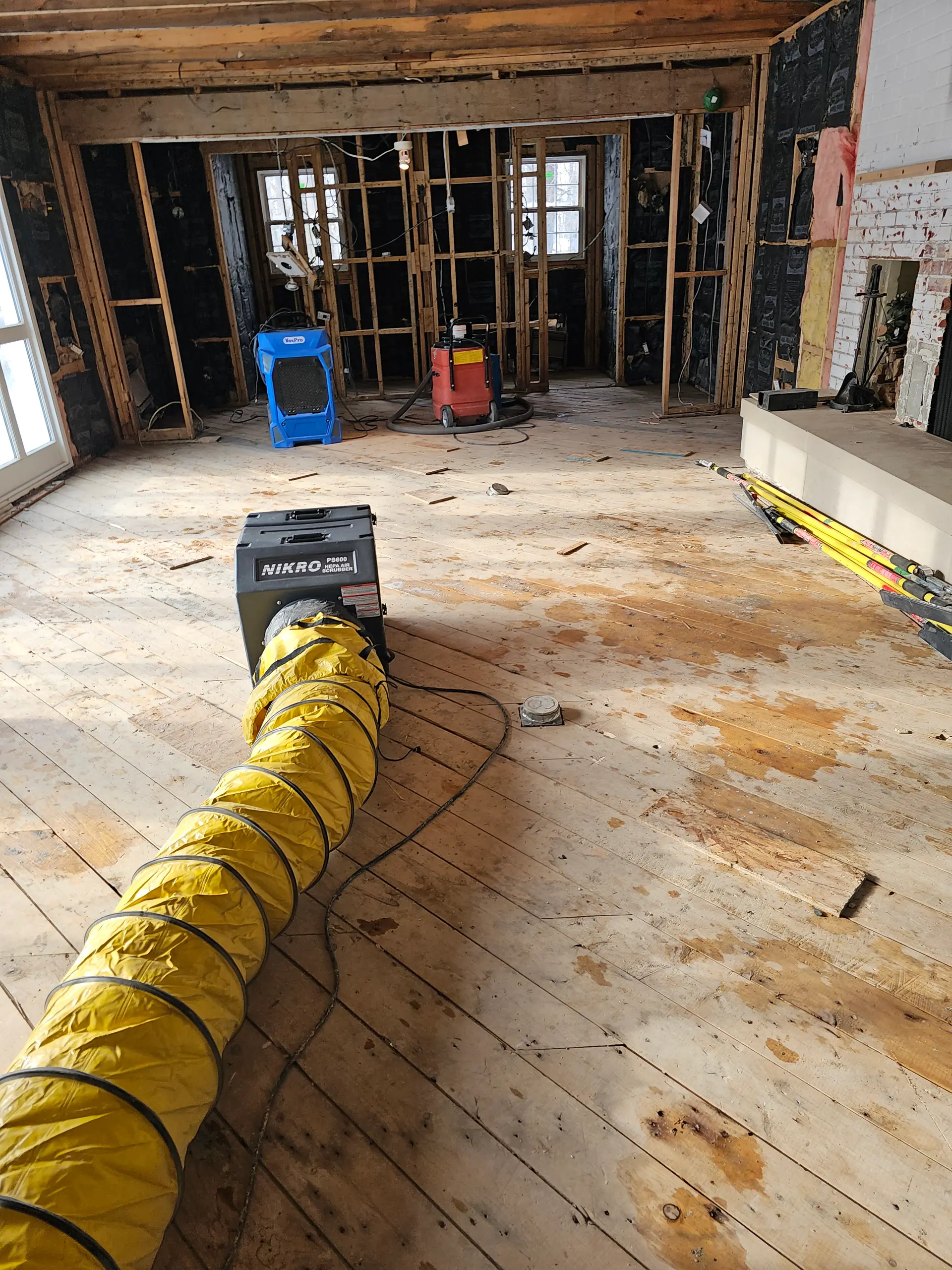 Pièce en rénovation après sinistre avec murs ouverts, plancher de bois exposé, déshumidificateur et gaine de ventilation pour l’assèchement.