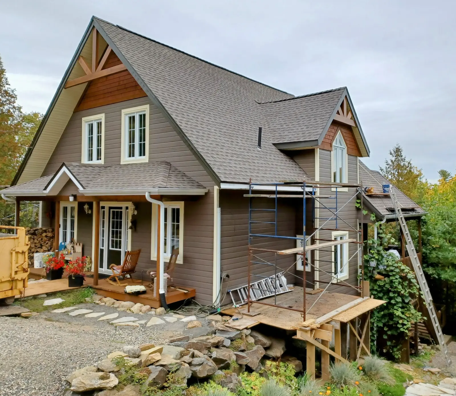 Gable-roof house under renovation with scaffolding, brown siding and shingle roofing, in a wooded setting