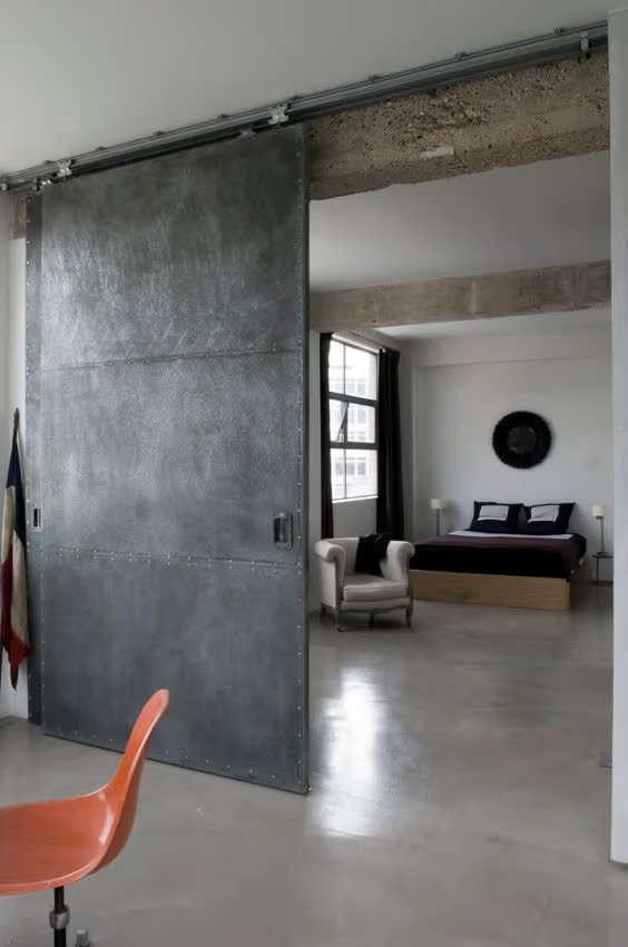 Porte coulissante industrielle en métal gris sur rail apparent, séparant un loft moderne avec béton exposé et chambre ouverte.
