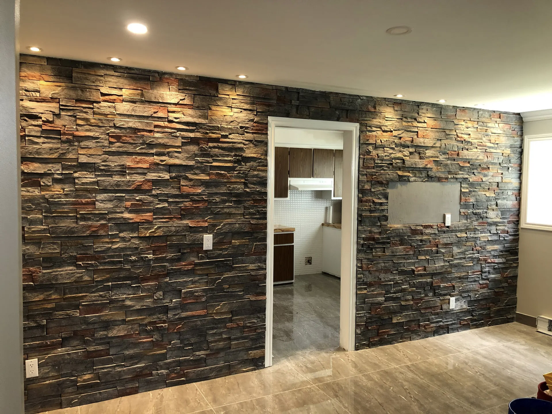 Interior accent wall clad in dark stacked stone, illuminated by recessed spotlights, with an opening leading to a modern kitchen.
