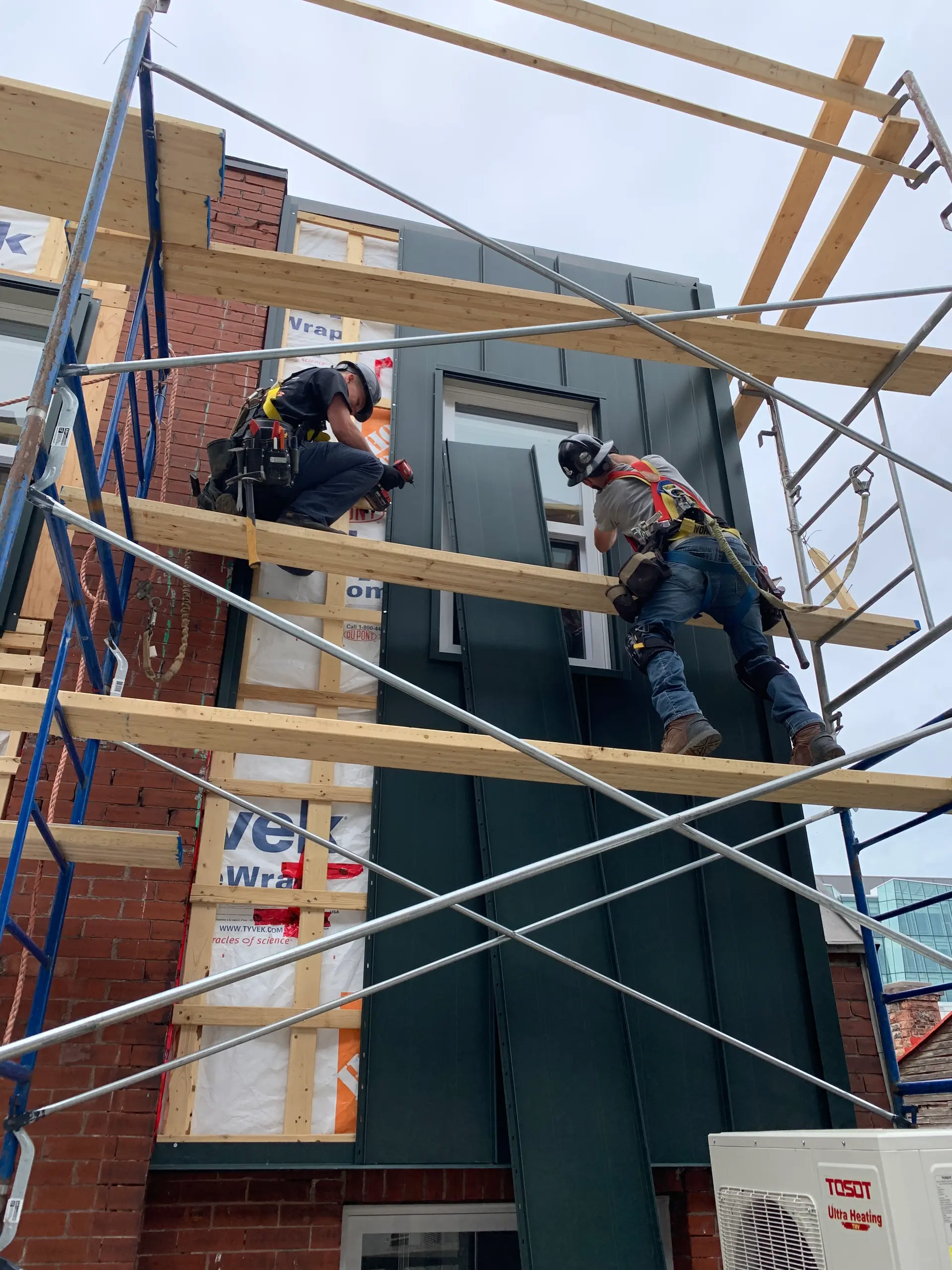 Construction workers installing vertical metal siding on a building facade using scaffolding