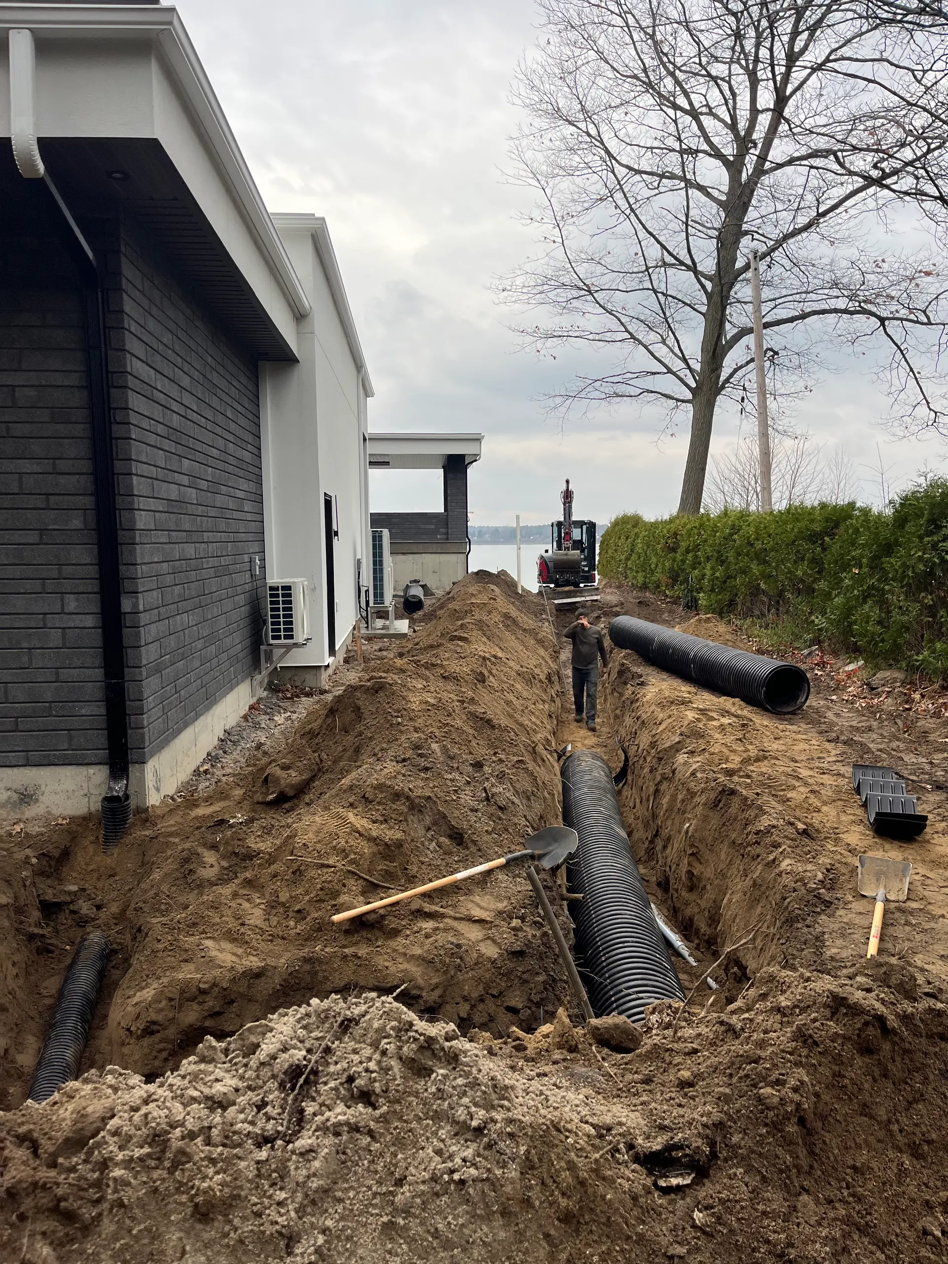 Chantier d’excavation résidentielle avec tranchées profondes, tuyaux de drainage noirs, pelle mécanique et installation de drainage extérieur le long d’une maison moderne