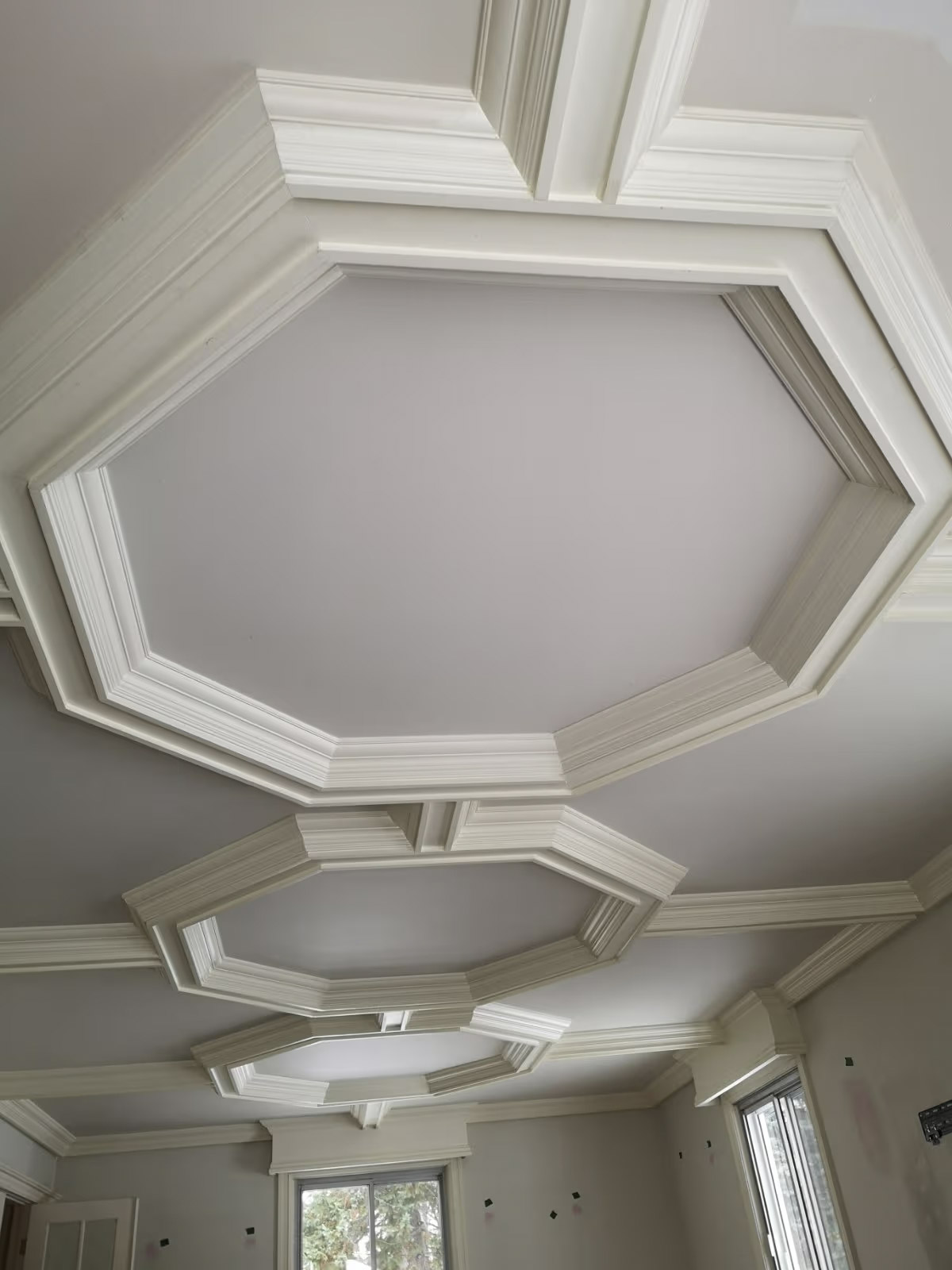 Plafond décoratif élégant avec caissons octogonaux, moulures blanches superposées et finition gris pâle dans une pièce lumineuse.