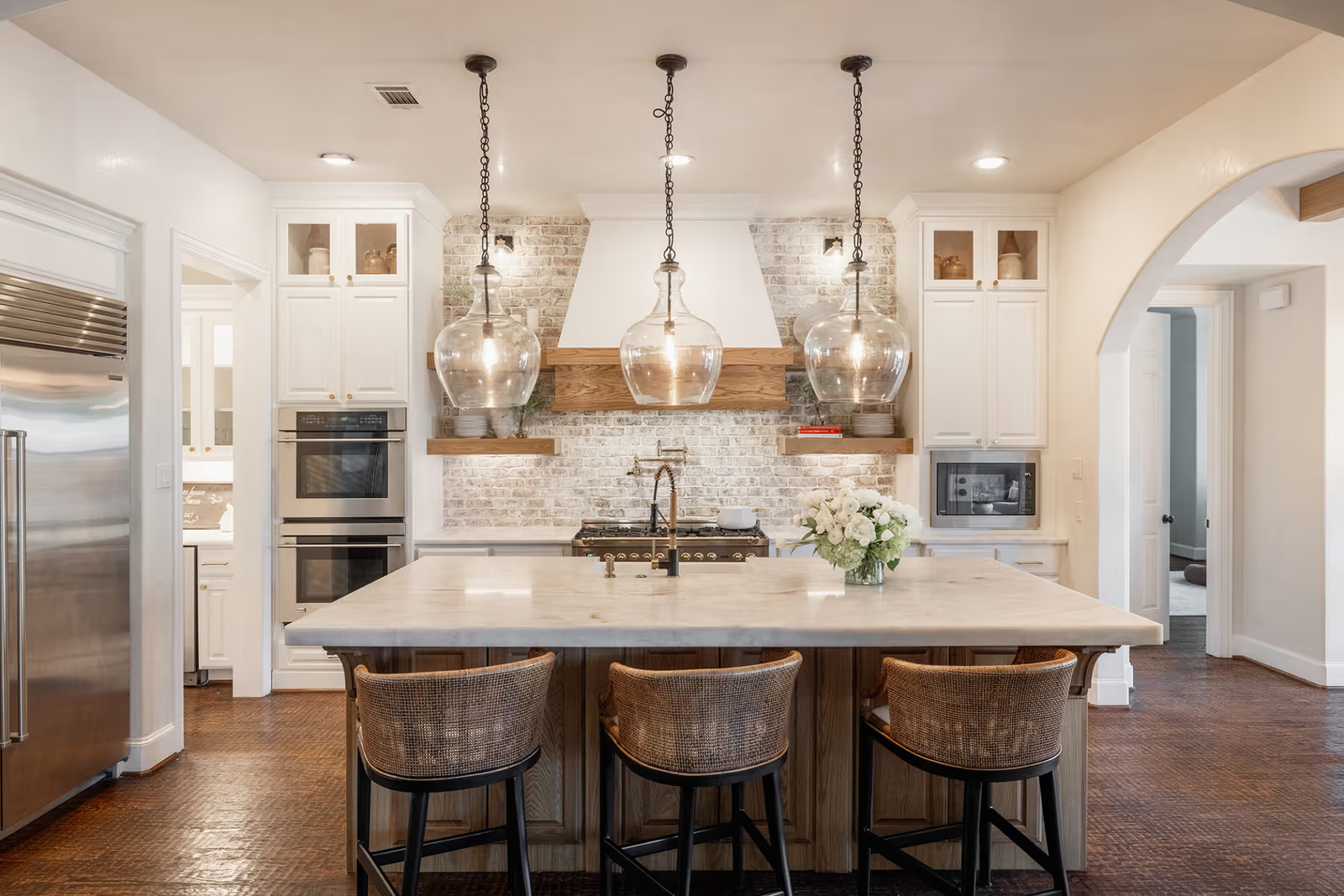 Cuisine élégante avec grand îlot central en bois, comptoir clair, trois luminaires suspendus en verre, armoires blanches, dosseret de brique et électroménagers en acier inoxydable.
