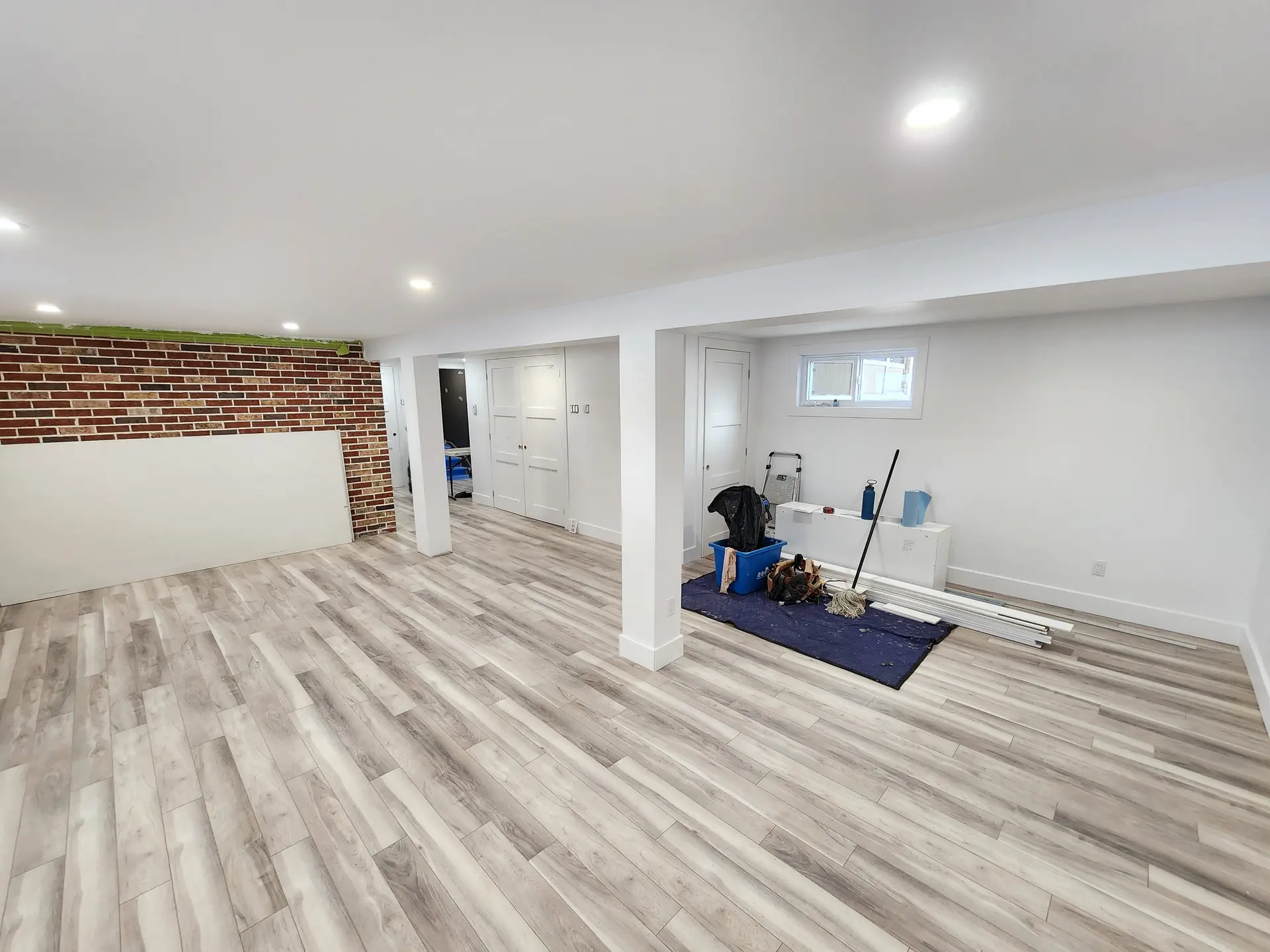 Sous-sol rénové lumineux avec plancher imitation bois, murs blancs, éclairage encastré, portes intérieures et mur de briques décoratif