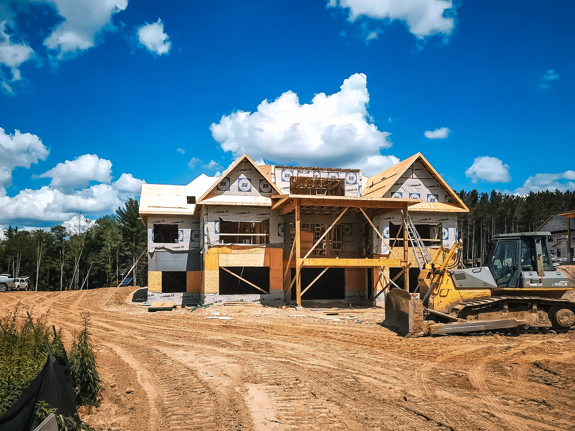 Two-story house under construction on sandy lot with Tyvek weatherproofing, wooden scaffolding and heavy machinery on site.