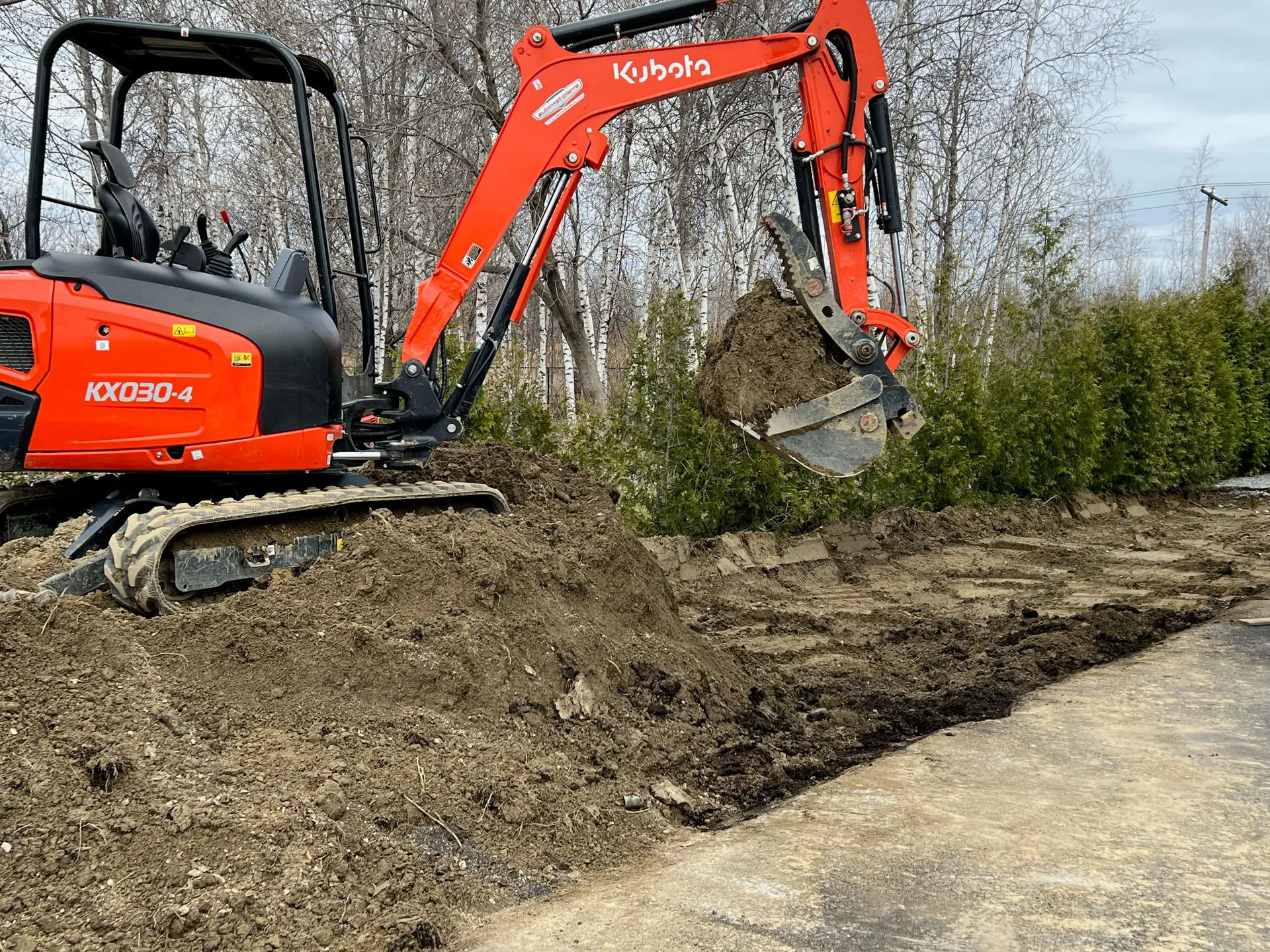 Mini-pelle Kubota orange en action sur un chantier d’excavation, creusant la terre avec son godet hydraulique devant une haie d’arbres.