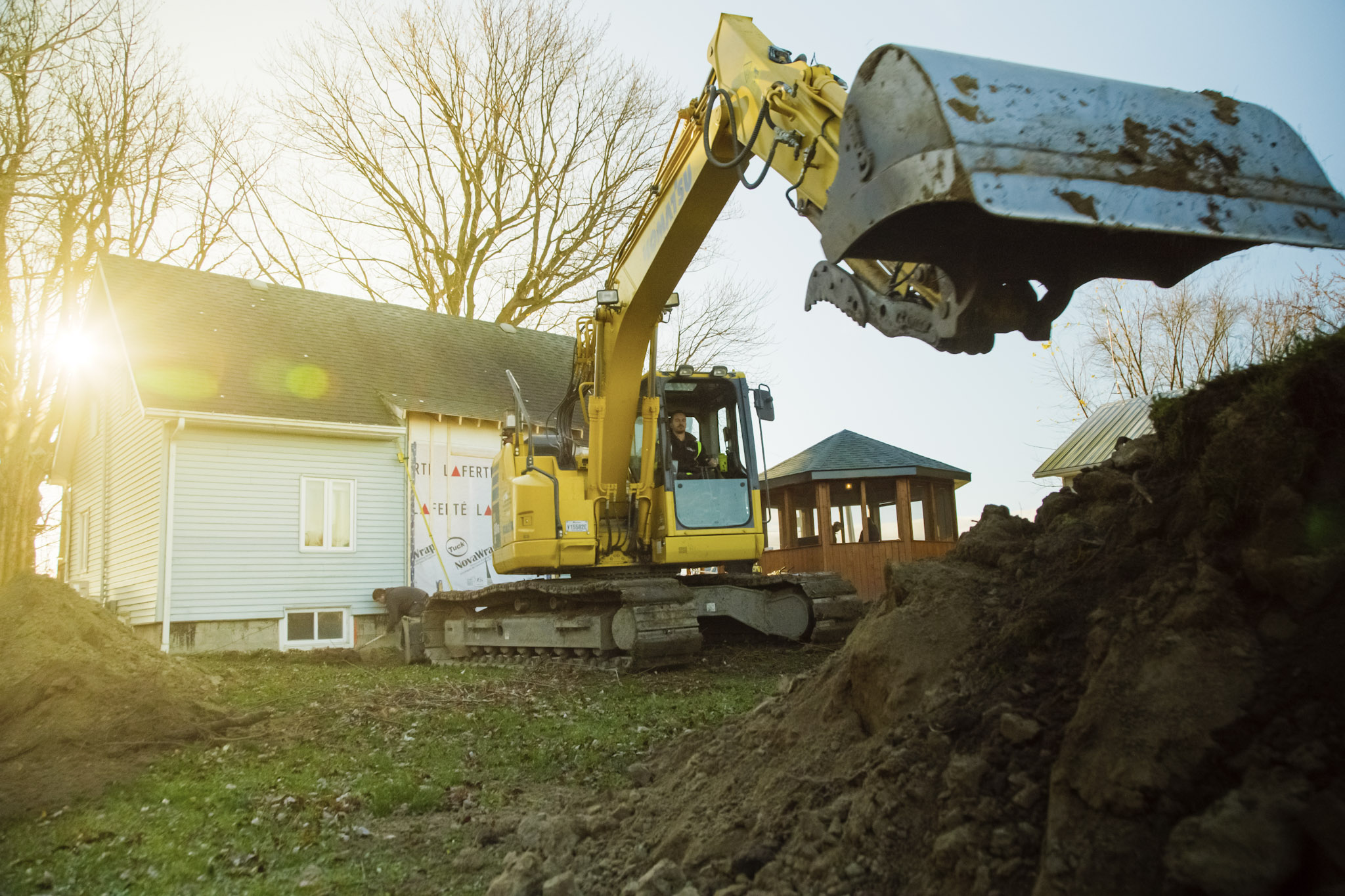 Excavatrice jaune travaillant près d’une maison résidentielle avec godet chargé de terre, piles de sol, arbres et lumière de fin de journée.