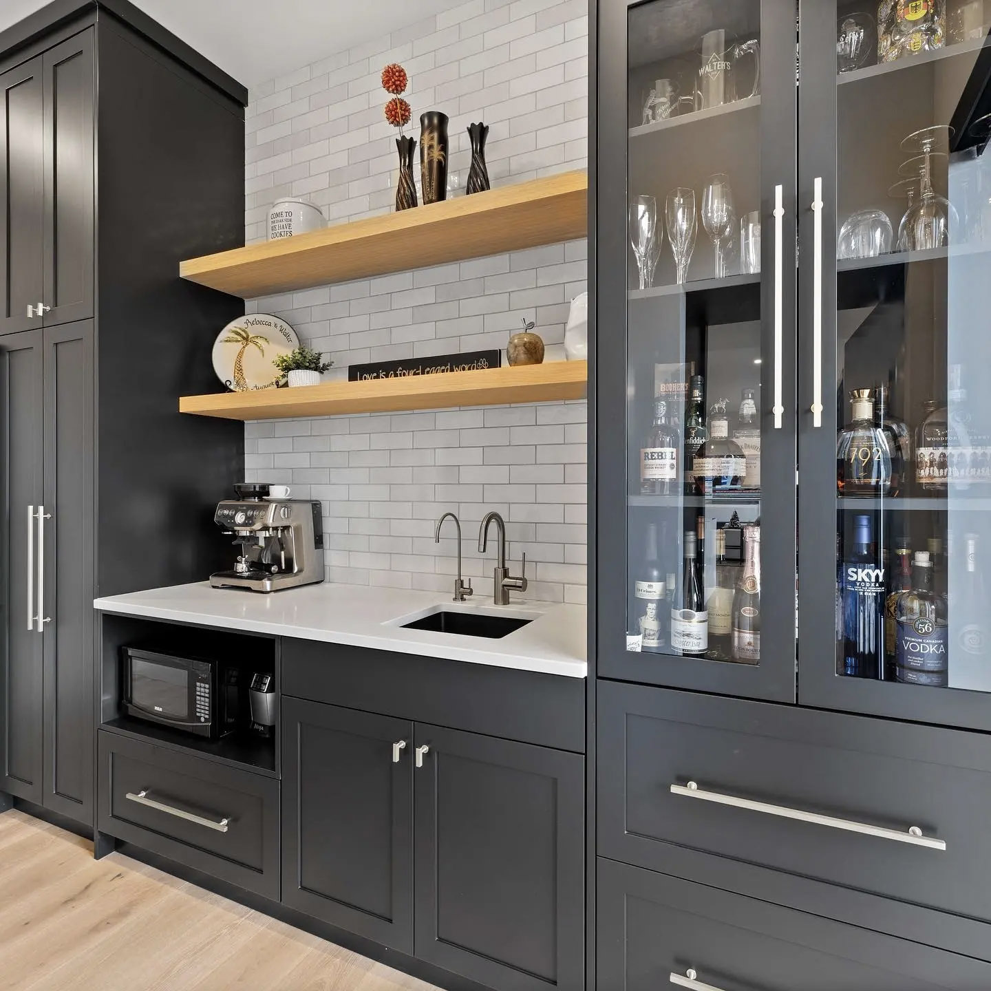 Black kitchen bar with a sink, espresso machine, wood shelves, and a glass-front cabinet for bottles and glassware.