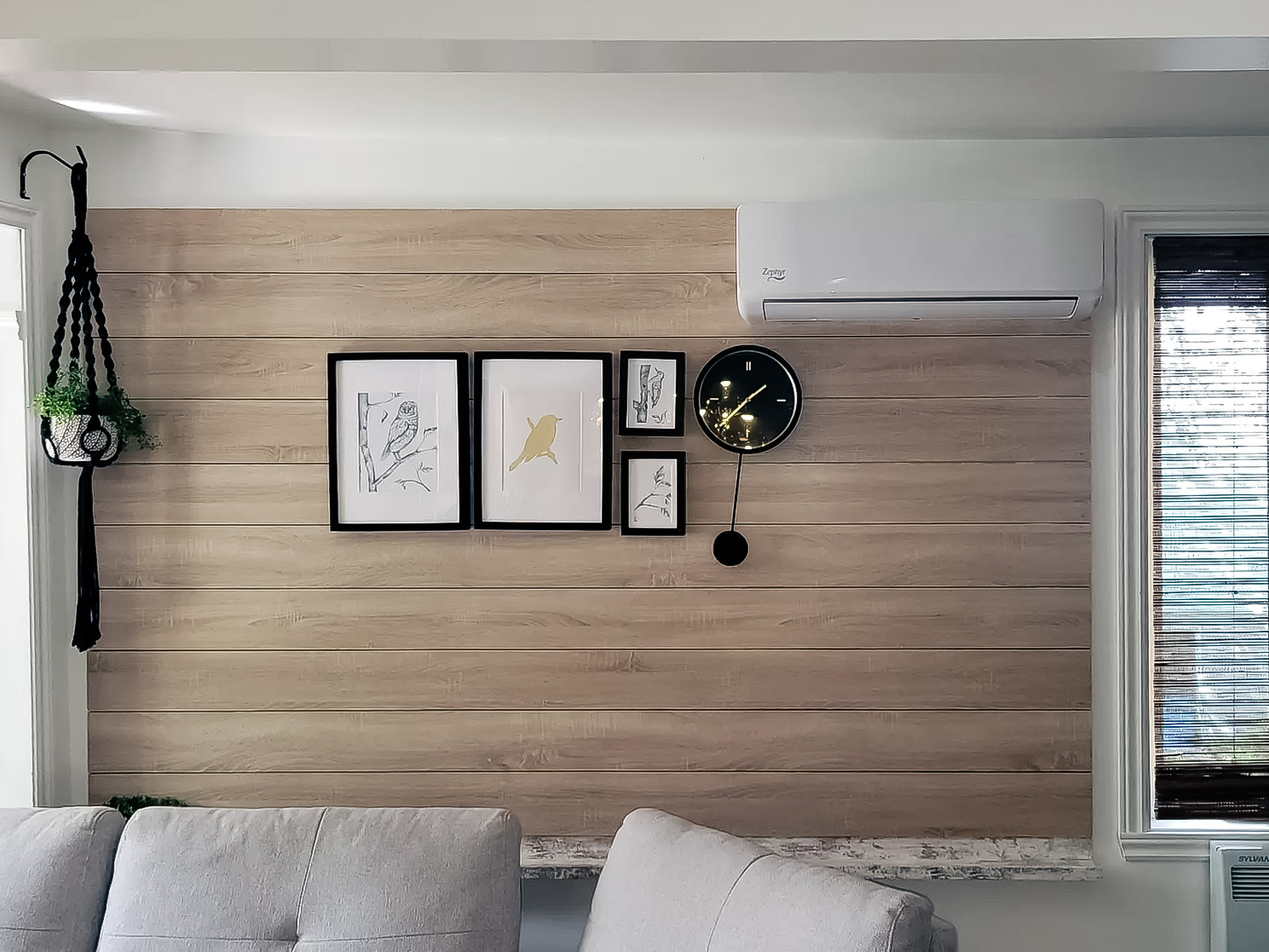 Light wood wall with wall-mounted air conditioning, decorative frame gallery, black designer clock and hanging plant in a modern living room.