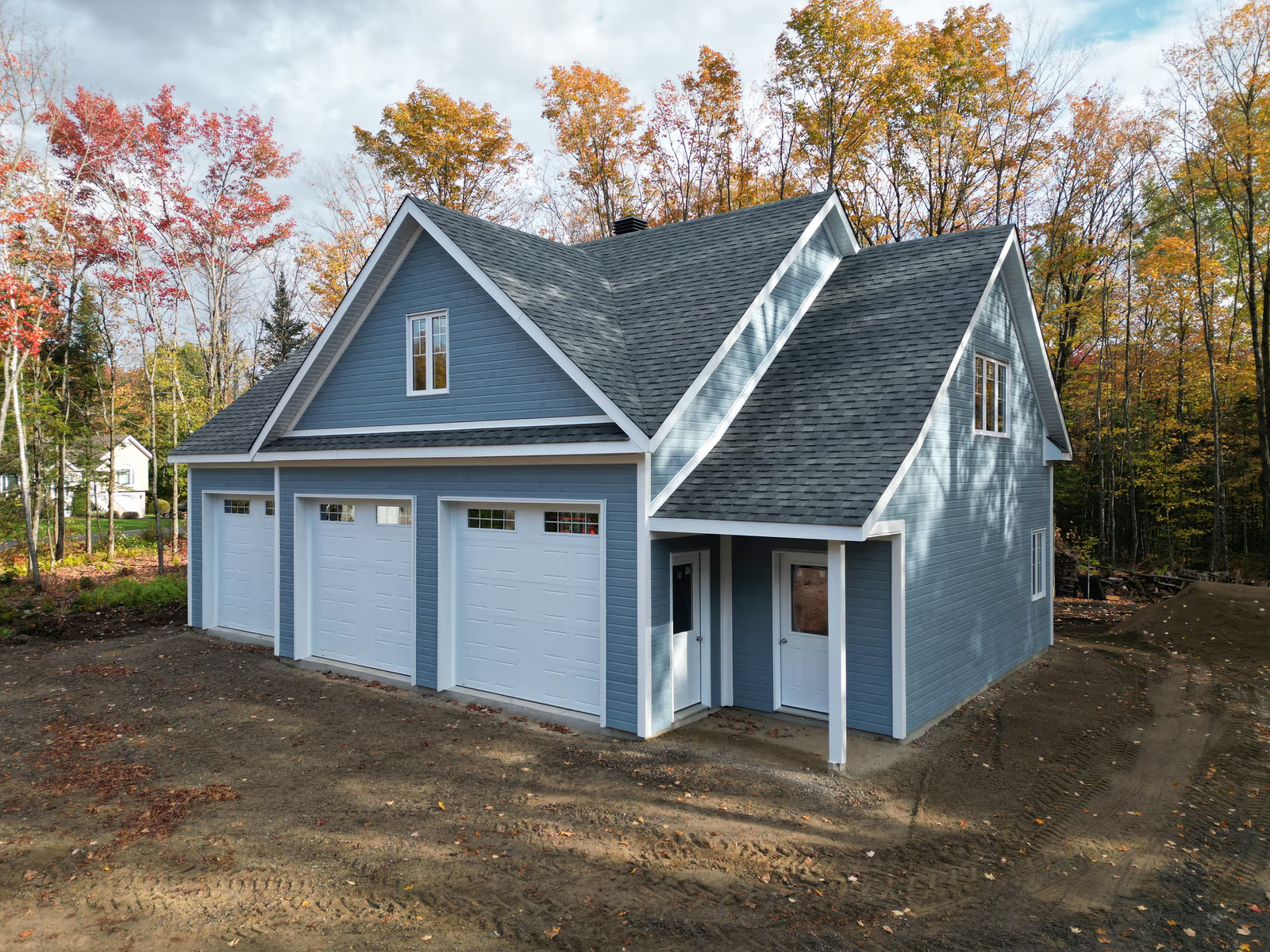Blue residential garage with three white doors, shingle roof, horizontal exterior siding, windows, entry doors and construction yard surrounded by autumn trees