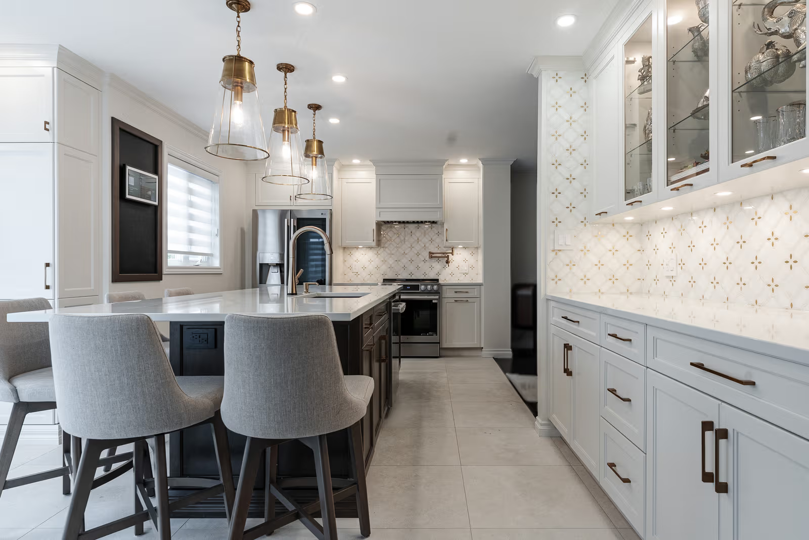 Bright elegant kitchen with white cabinetry, a large dark central island, light countertop, glass and brass pendant lights, illuminated display cabinets, and a decorative backsplash.