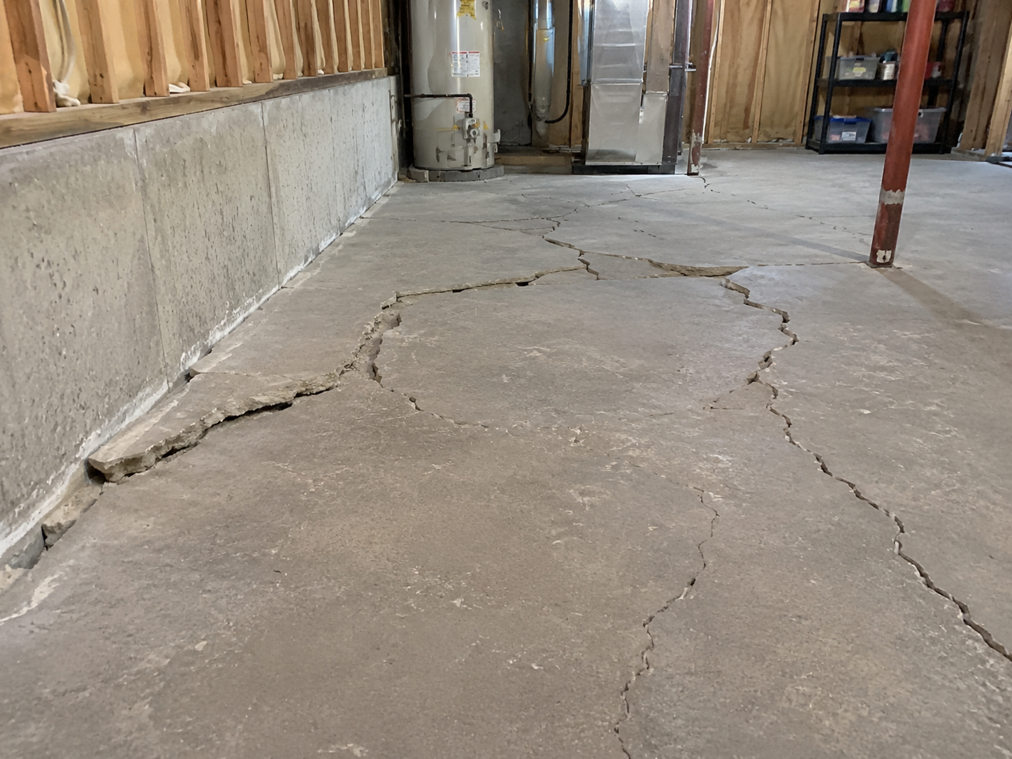 Un sous-sol non fini avec charpente apparente, équipements mécaniques et dalle de béton fortement fissurée et soulevée.