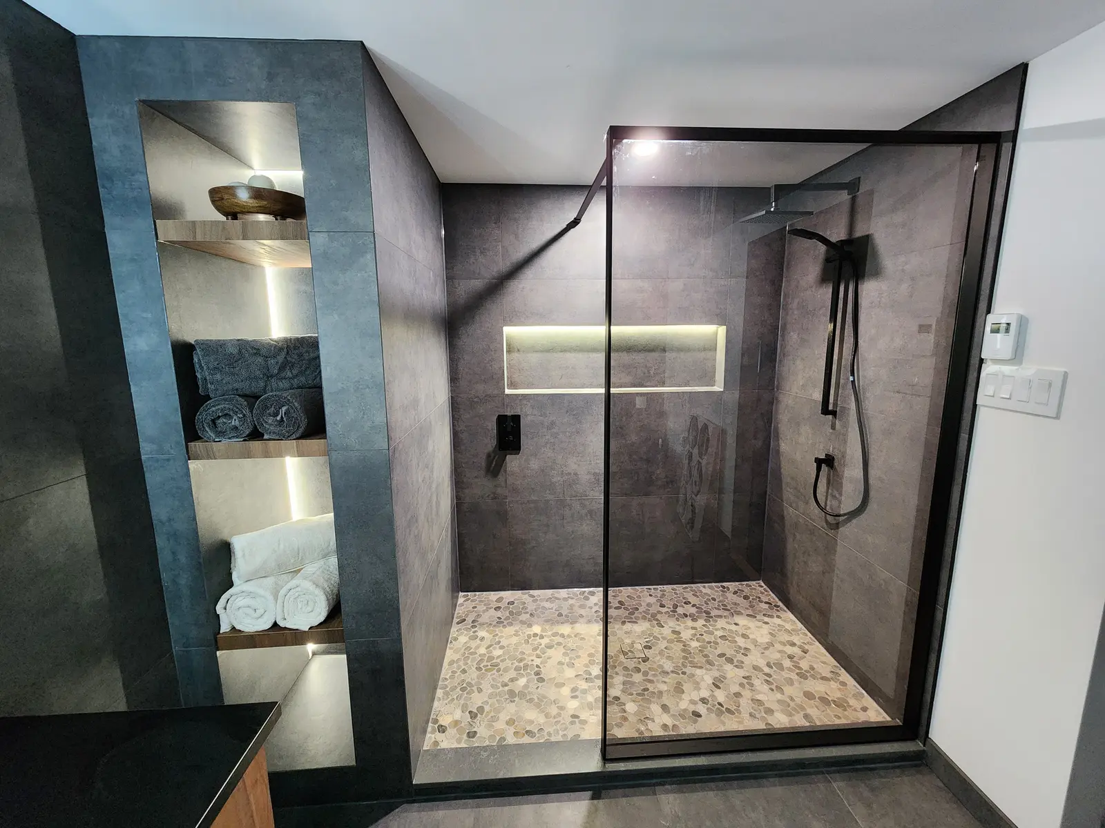 Douche italienne moderne avec paroi de verre noire, tuiles foncées, plancher en galets, niche murale éclairée et rangement intégré avec serviettes.