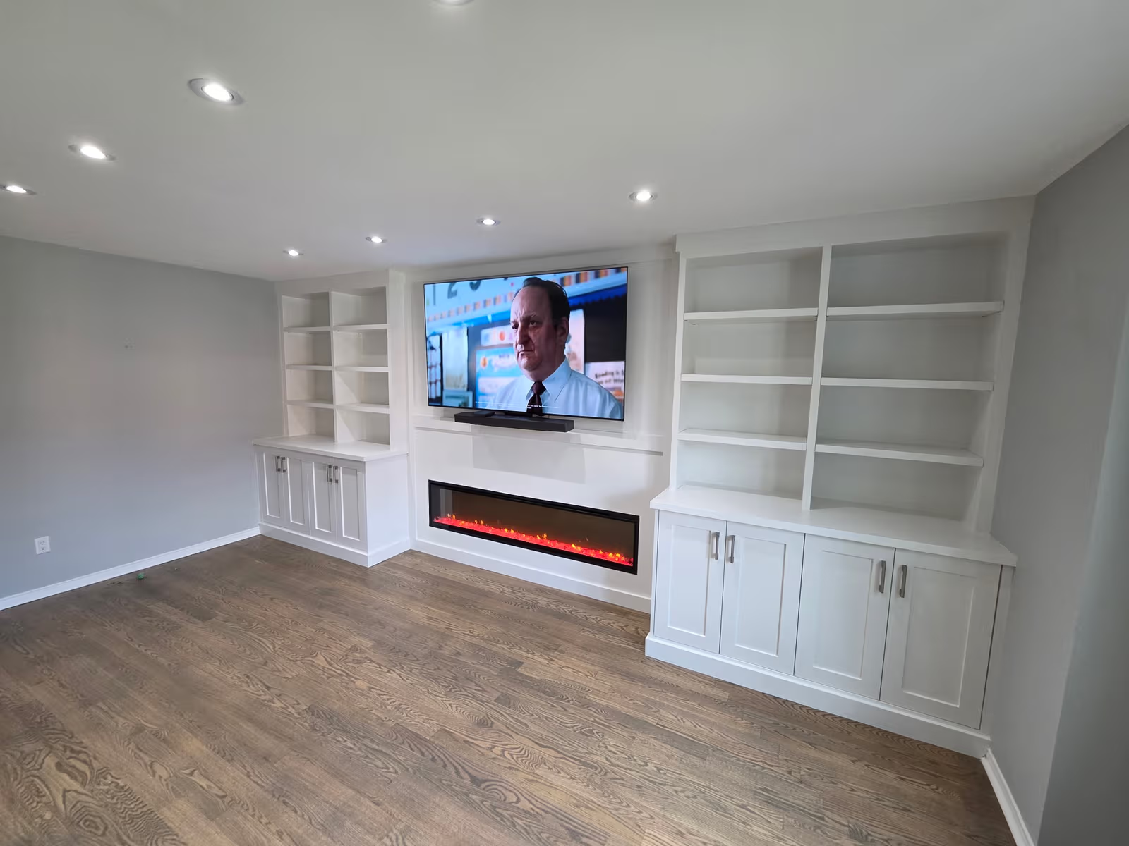 Renovated modern-style living room featuring a built-in electric fireplace under a wall-mounted TV, flanked by custom white bookshelves with lower cabinets and open shelving.