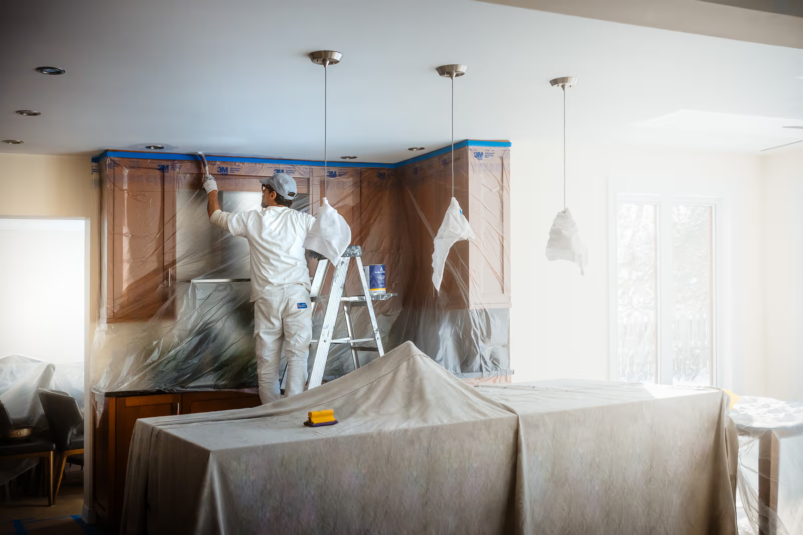 Worker preparing and painting a kitchen protected with plastic sheeting, with a step ladder, covered cabinetry, and wrapped pendant lights.