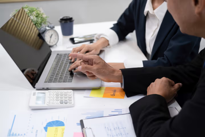 Two professionals collaborating on a laptop with calculator, financial charts and goal notes on a modern office desk.