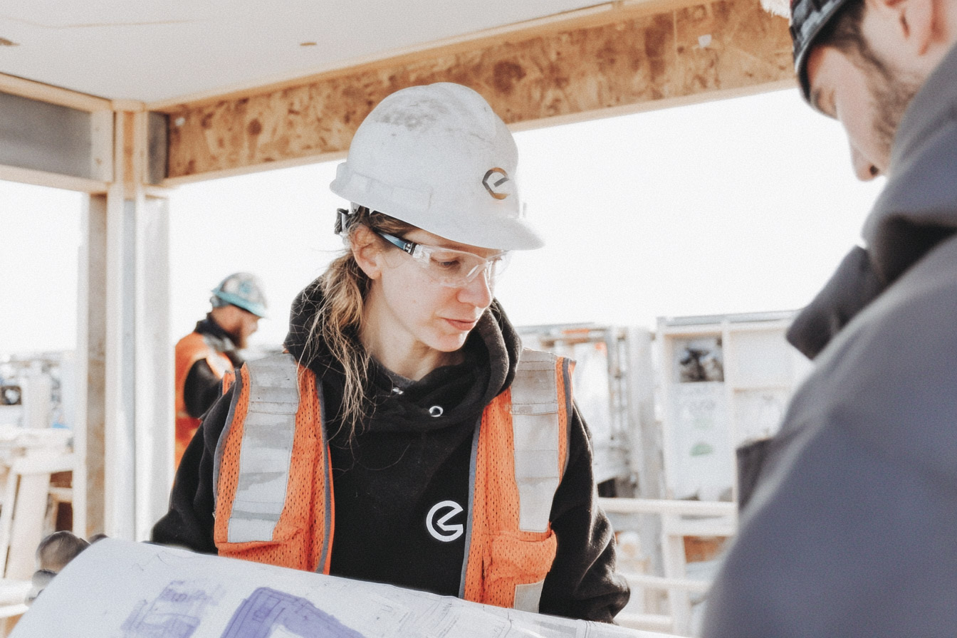 A construction worker wearing a hard hat, safety glasses, and a reflective vest reviews building plans with a colleague at a residential construction site.