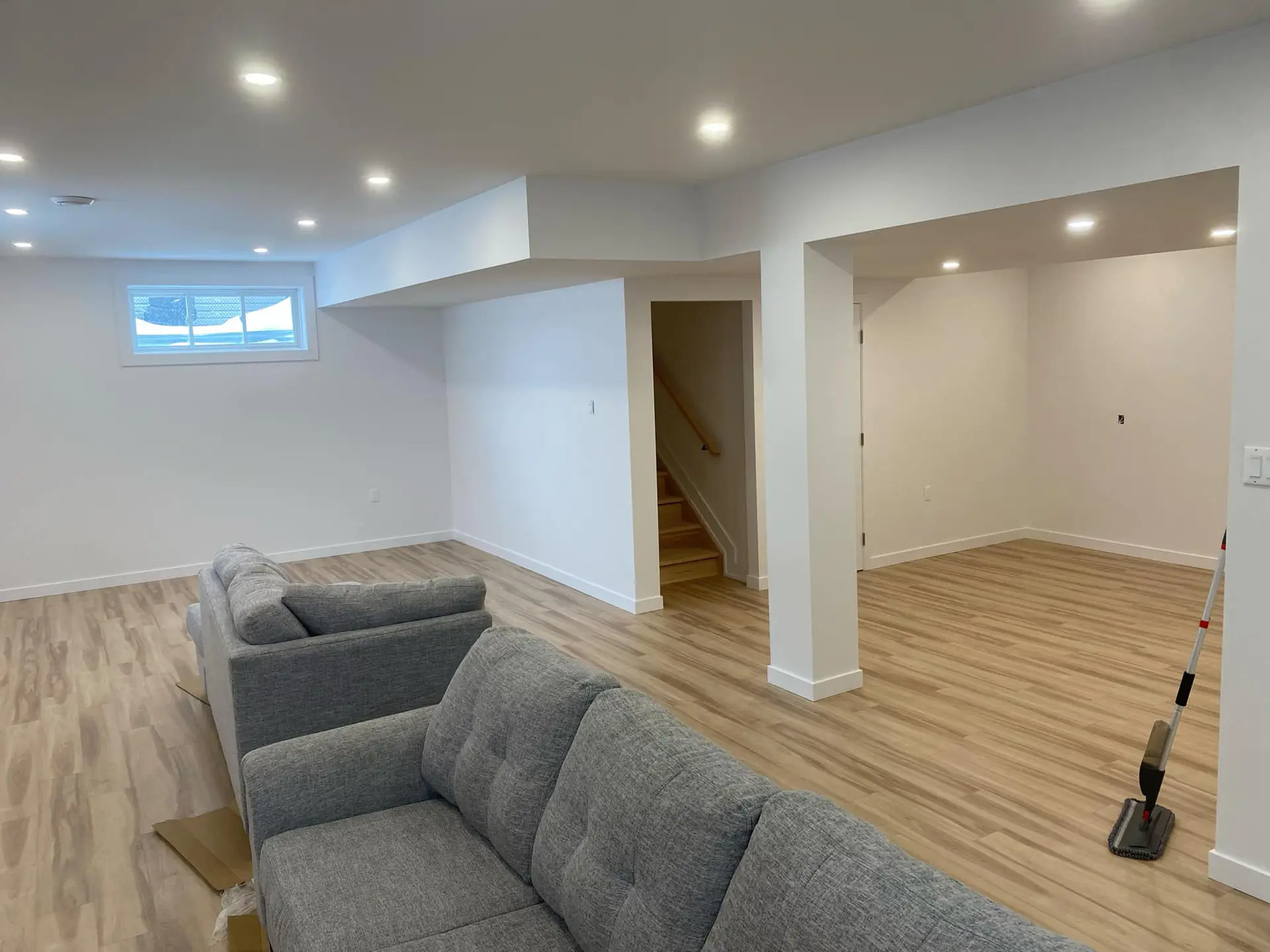 Renovated basement with light wood-look flooring, white walls, recessed lighting, grey sofa, and open multifunctional space.
