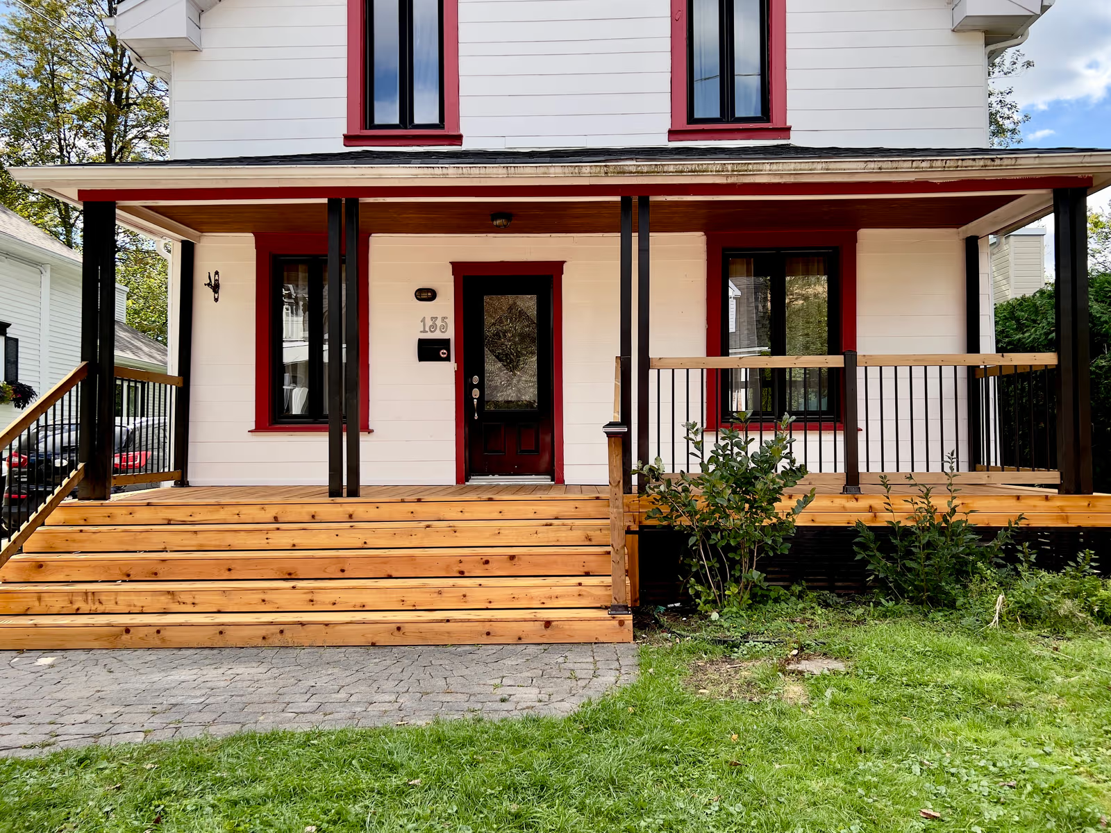 Maison rénovée avec galerie avant en bois, escalier en cèdre, rampes en métal noir, revêtement blanc et encadrements de fenêtres rouges