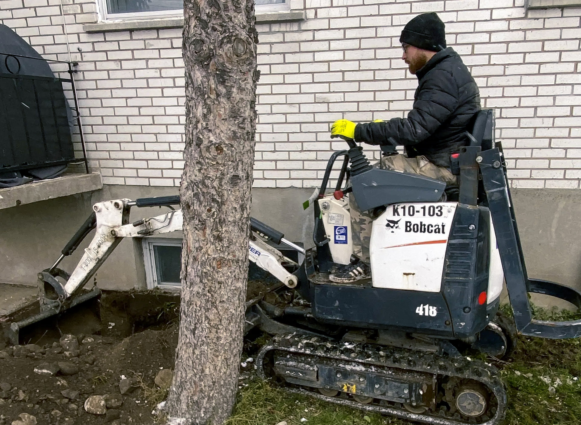 Worker operating a Bobcat mini excavator for excavation work near a white brick house