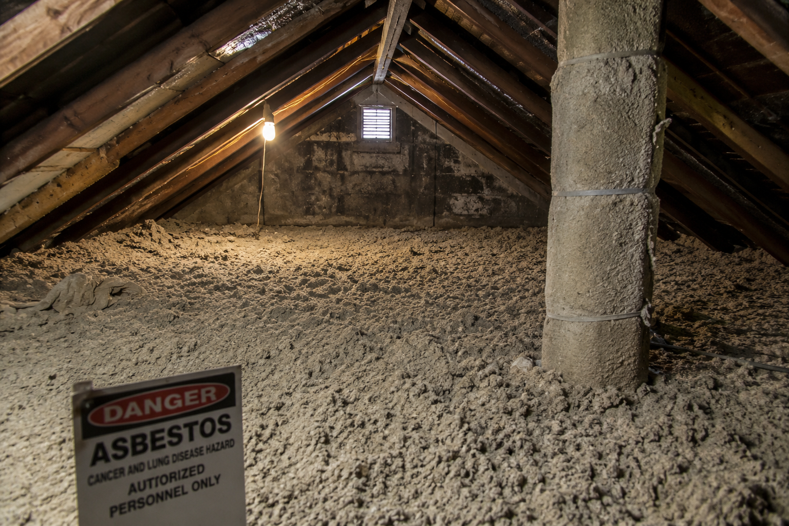 Old attic containing loose-fill insulation potentially contaminated with asbestos, with wooden framing, a vertical duct, and a hazard warning sign.