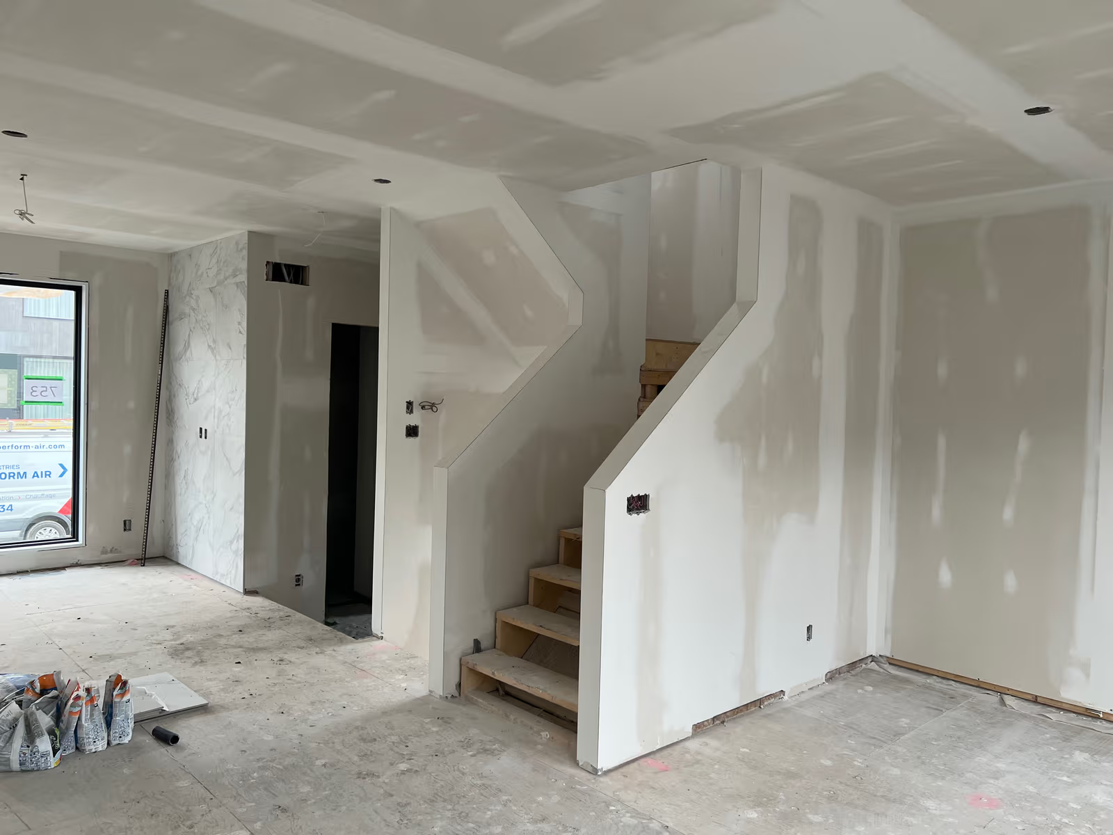 <br>House interior under renovation with drywall walls and ceiling, taped joints, a central staircase, and electrical openings before finishing work.