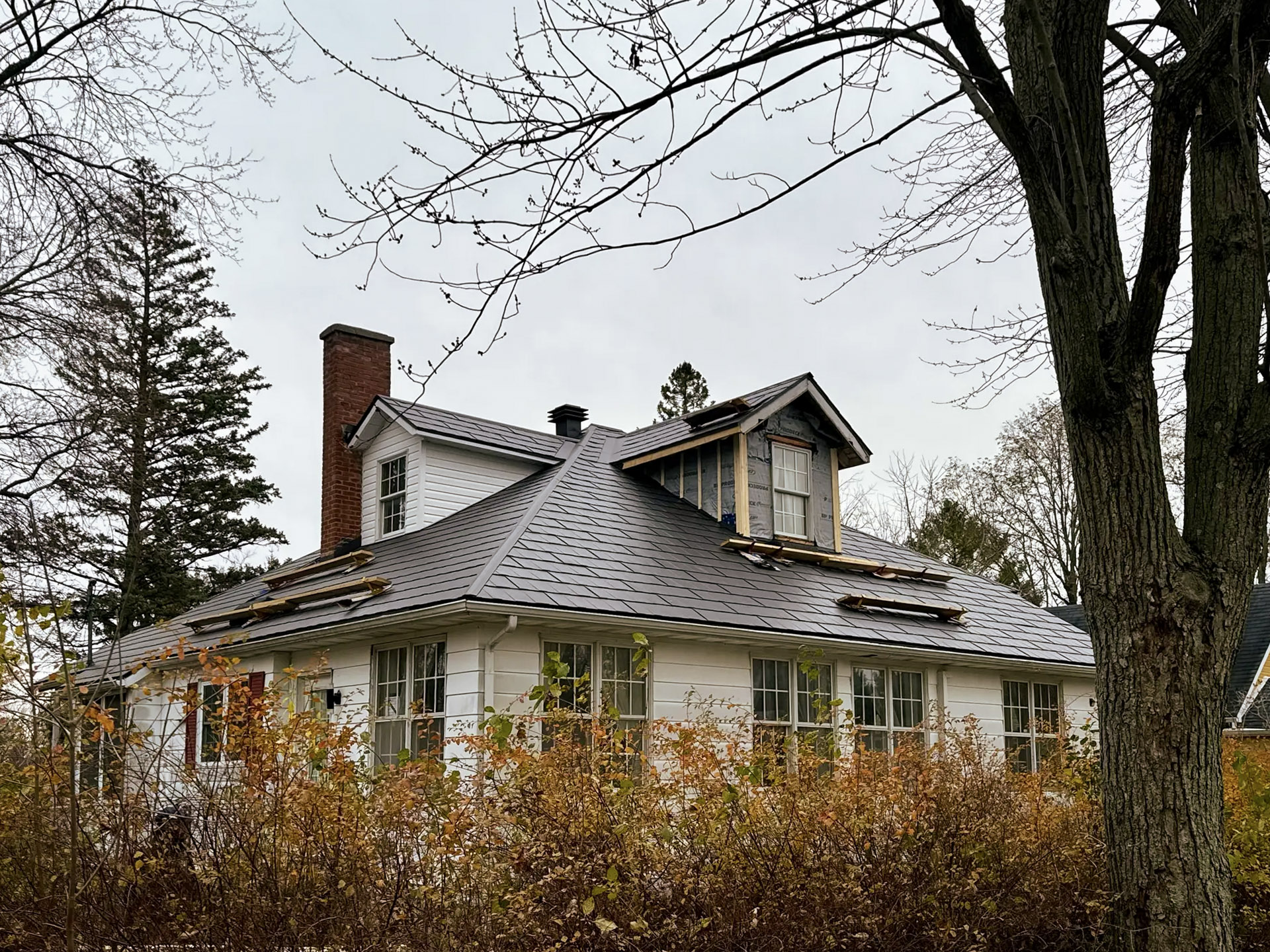 White residential house with dark metal roofing under renovation, dormer windows and brick chimney.