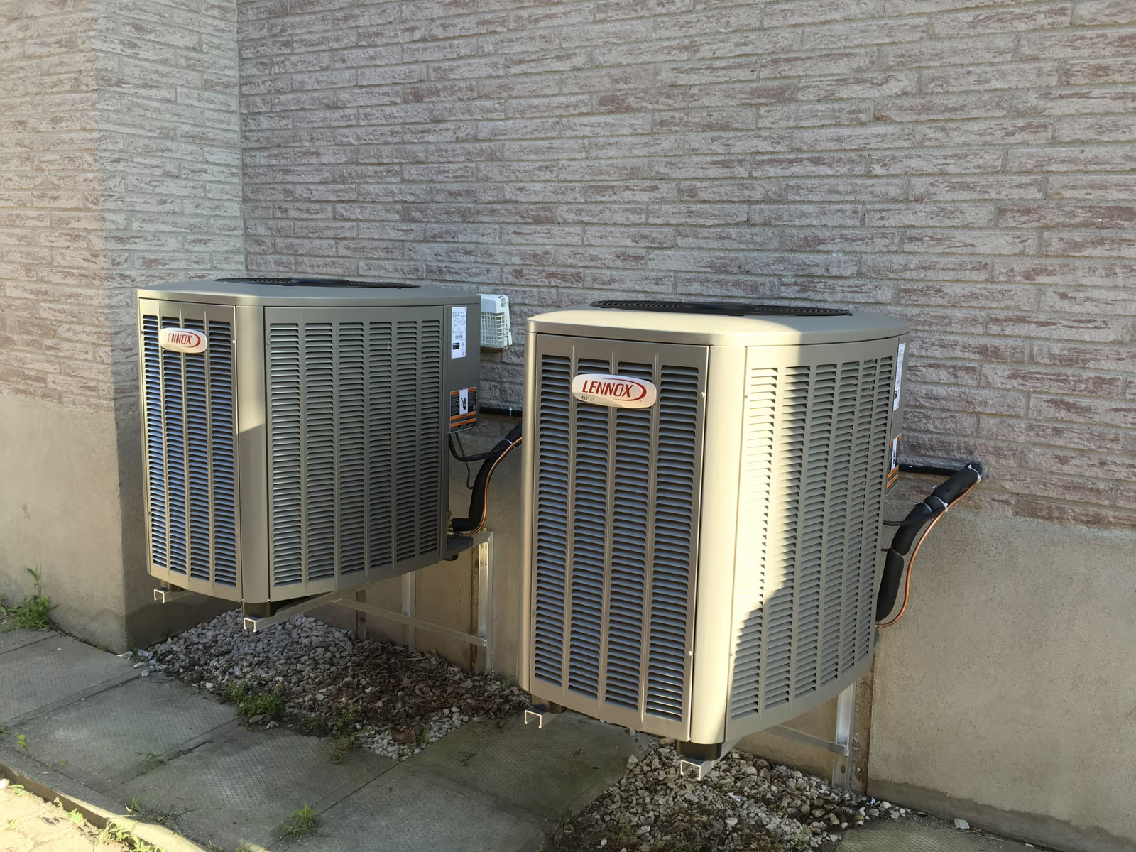 Climatiseurs extérieurs Lennox installés sur supports muraux contre un mur de brique d’une maison, système de climatisation résidentiel avec conduites et câblage visibles pour la ventilation et le refroidissement de l’habitation.