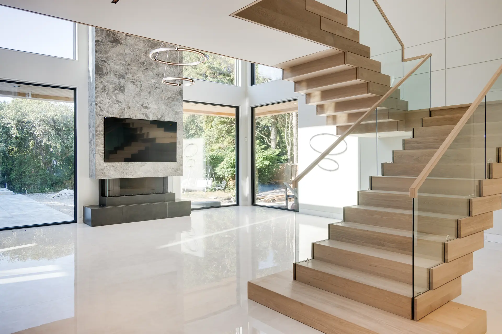 Escalier en bois moderne avec garde-corps en verre, dans un intérieur lumineux avec sol en carrelage poli, grandes fenêtres et cheminée en pierre contemporaine. 