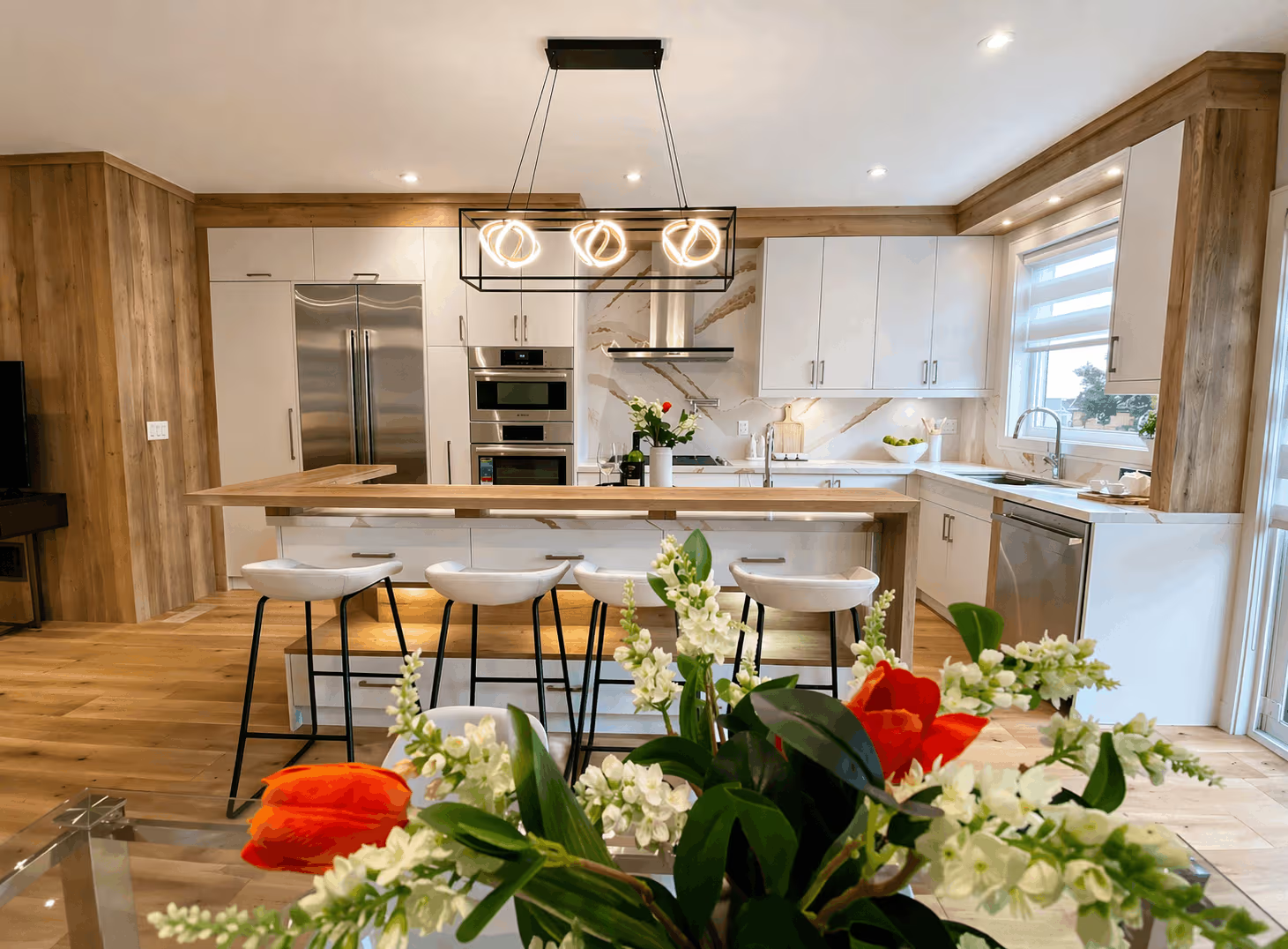 Cuisine moderne et lumineuse avec armoires blanches et bois naturel, grand îlot avec quatre tabourets, électroménagers en acier inoxydable, dosseret effet marbre et suspension contemporaine.