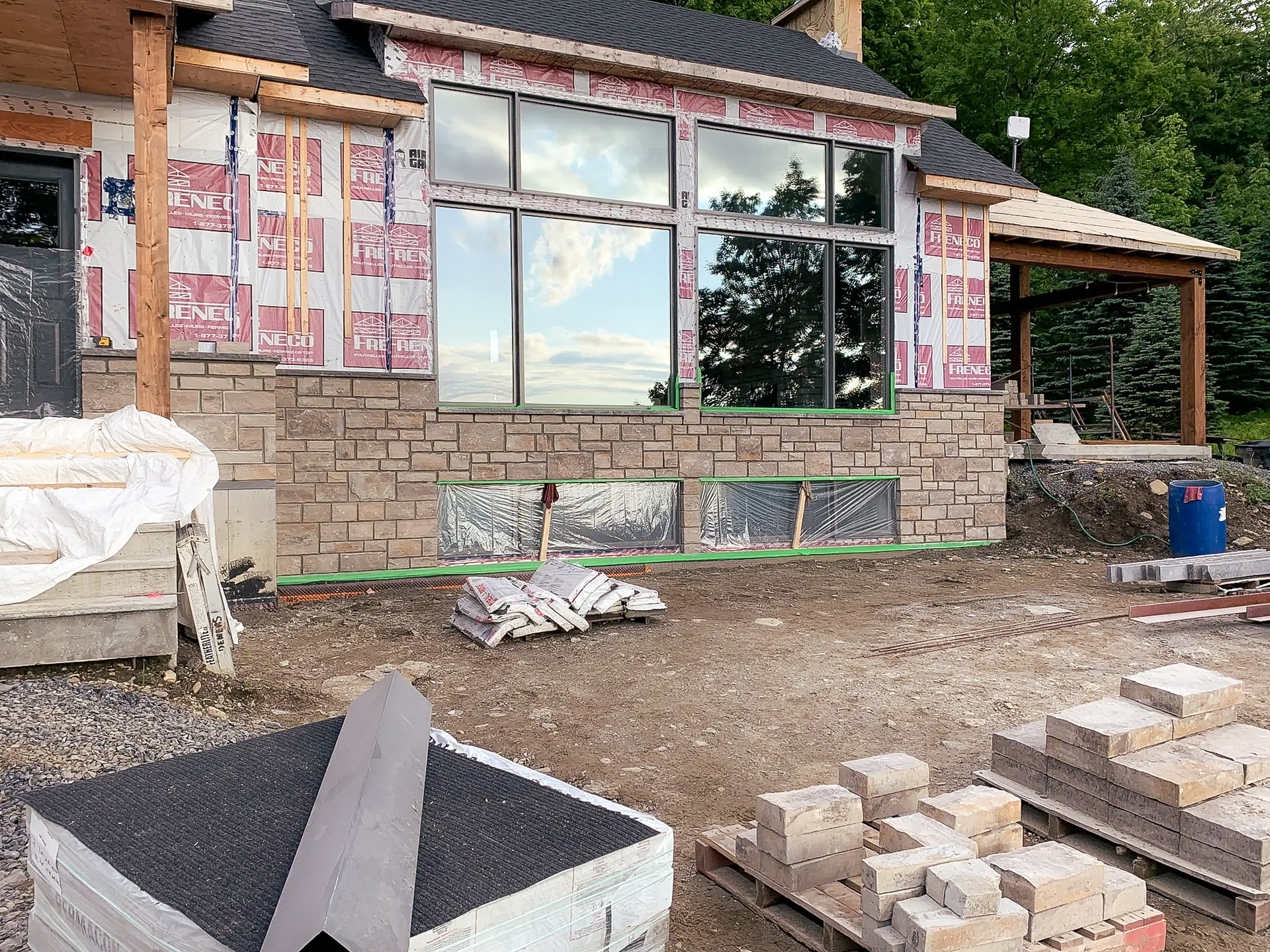House under construction with partially installed stone siding, large windows, and construction materials on the ground.