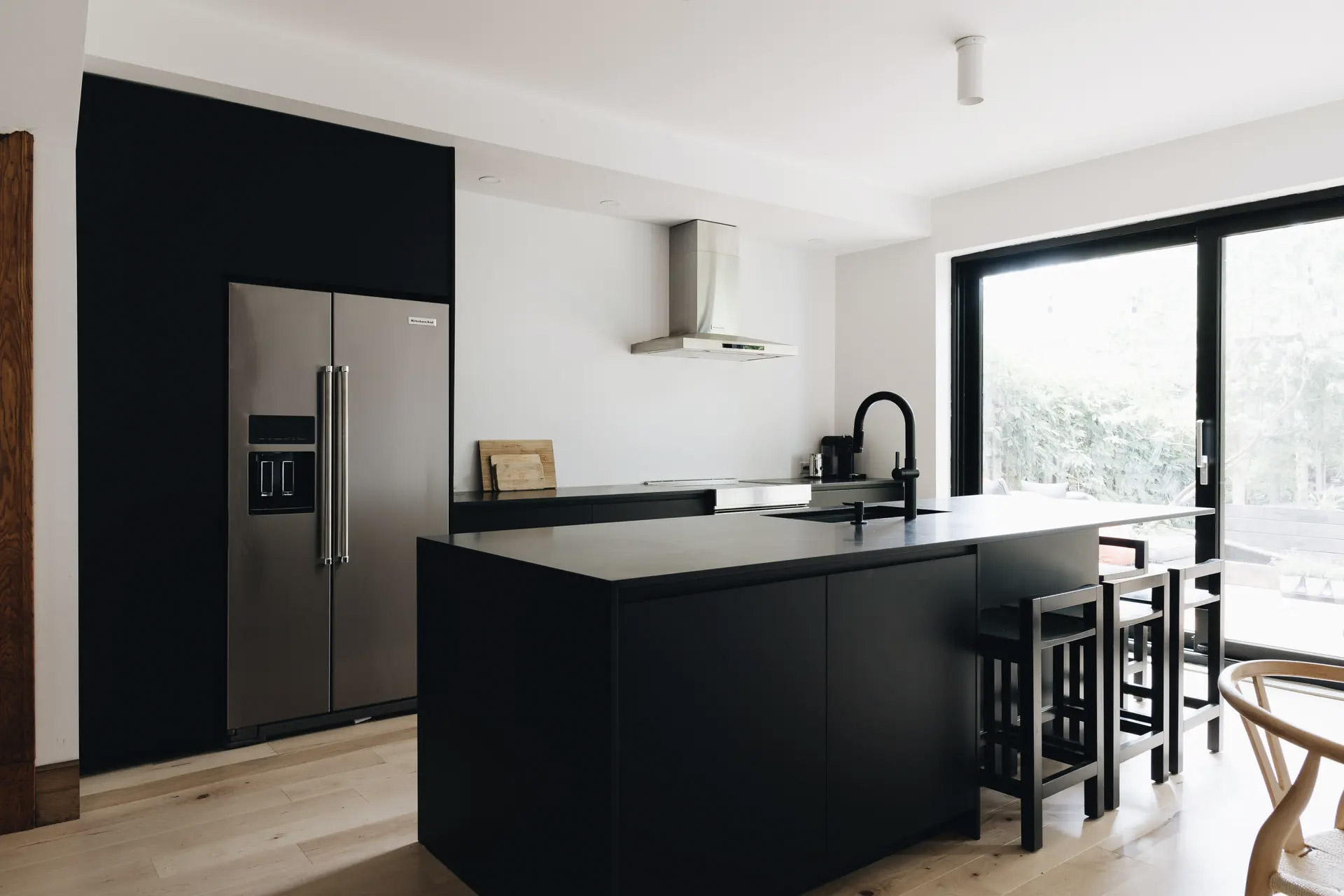 Cuisine noire contemporaine avec grand îlot central, comptoir minimaliste, réfrigérateur en acier inoxydable, robinet noir, hotte murale, tabourets, plancher en bois clair et grande porte-patio vitrée laissant entrer la lumière naturelle.