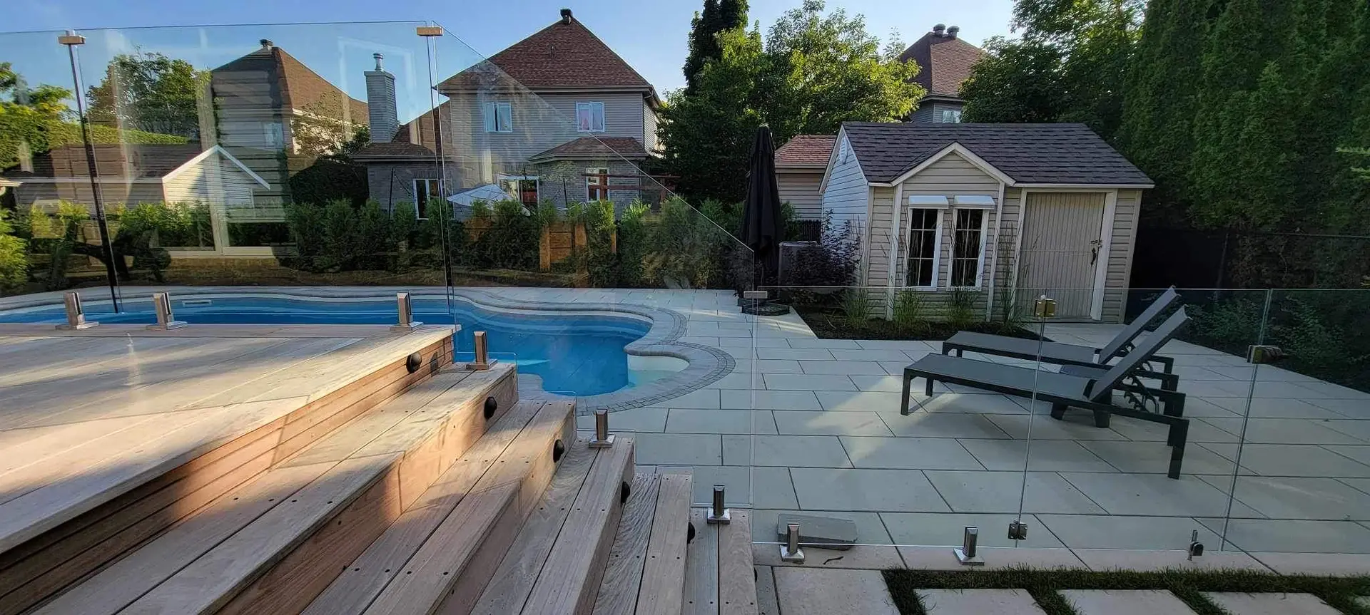 Backyard with inground pool, glass fence, raised wood terrace, patio slabs, garden shed, lounge chairs and mature hedges