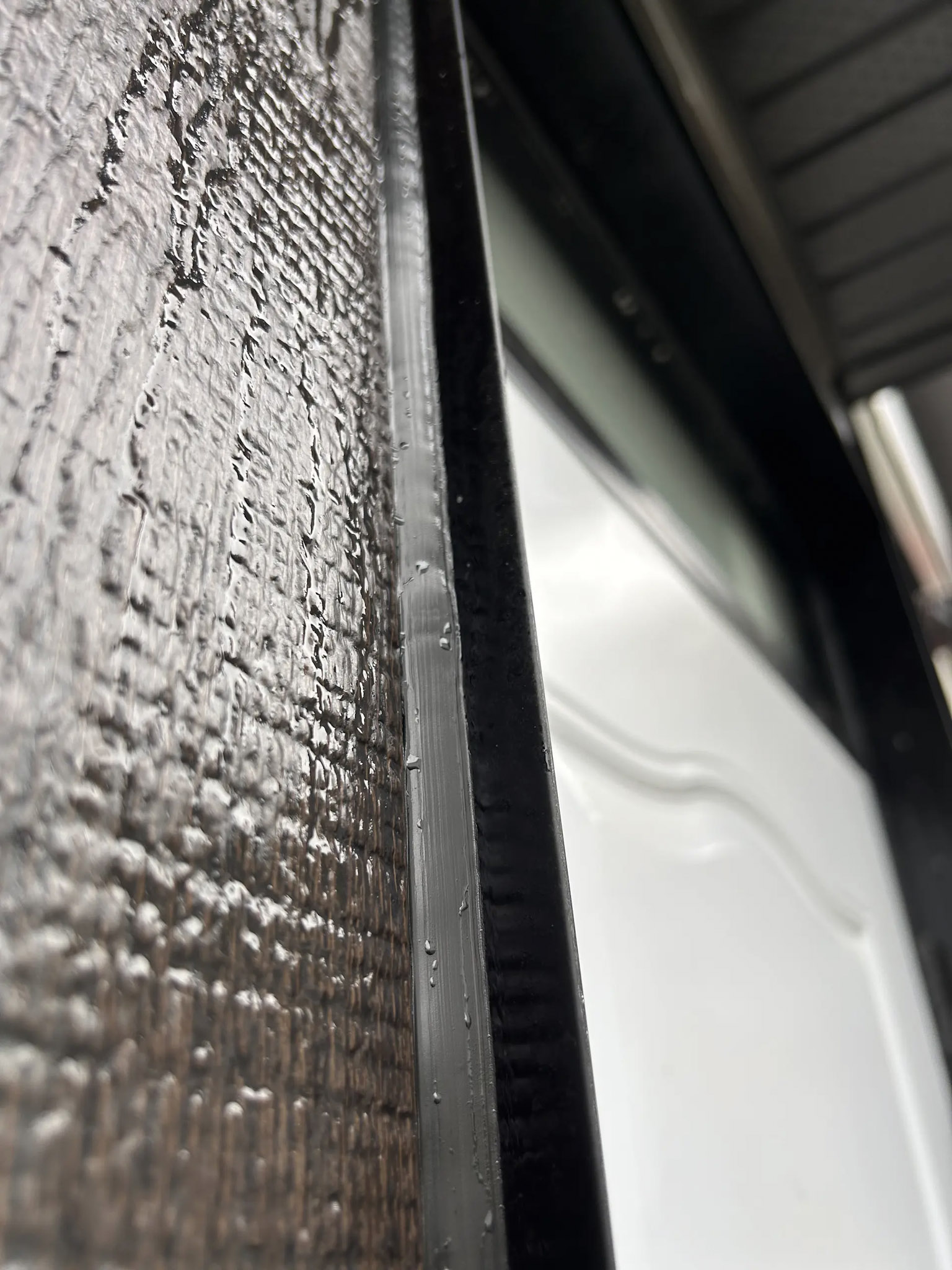 Close-up of a black weather seal between textured exterior siding and a white door, with visible moisture droplets.