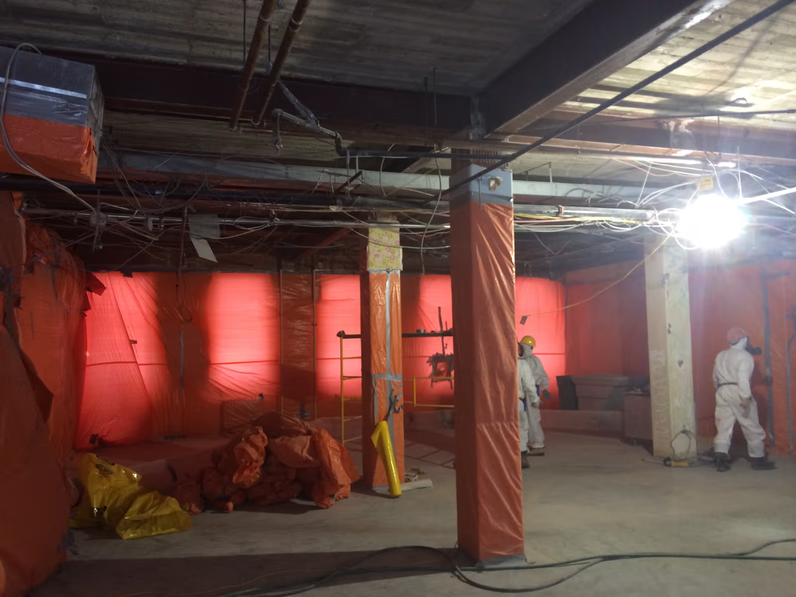 Chantier intérieur de décontamination avec confinement en polyéthylène orange, câblage apparent et travailleurs en combinaison de protection
