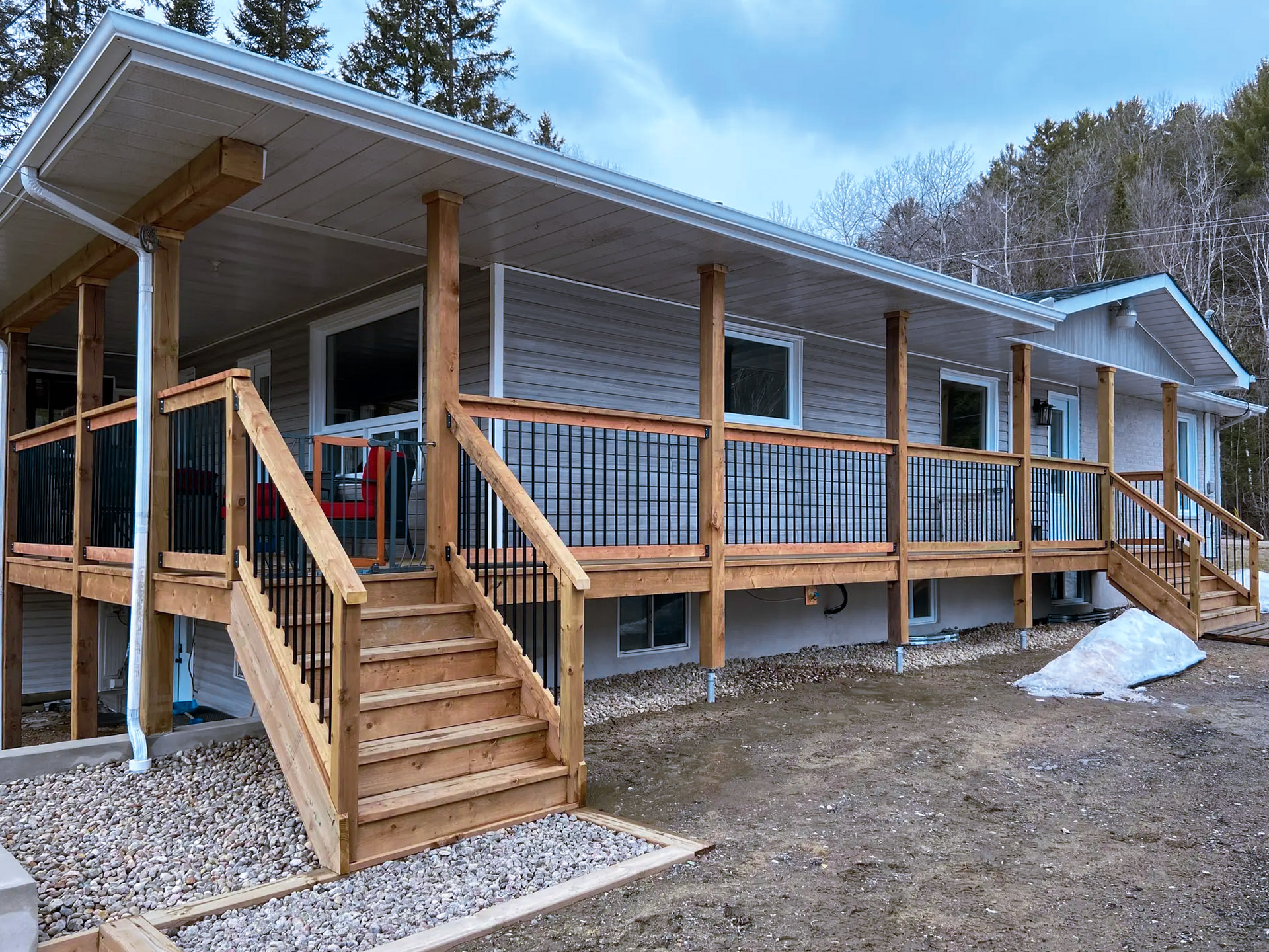 Grand patio en bois surélevé avec escaliers, rampes noires et poteaux de soutien devant une maison résidentielle