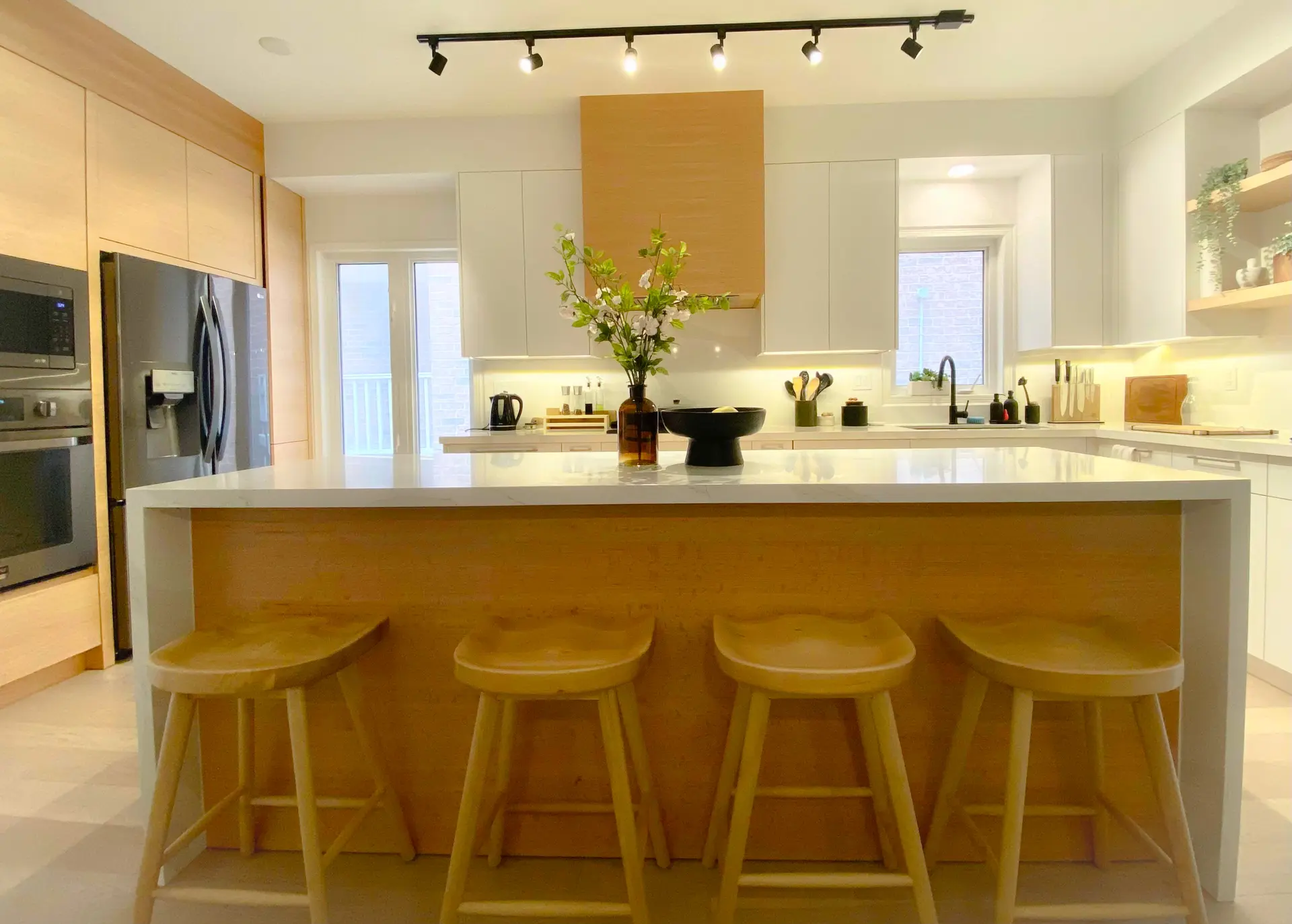 Bright modern kitchen with a large central island, white countertop, light wood cabinetry, four stools, and track lighting.