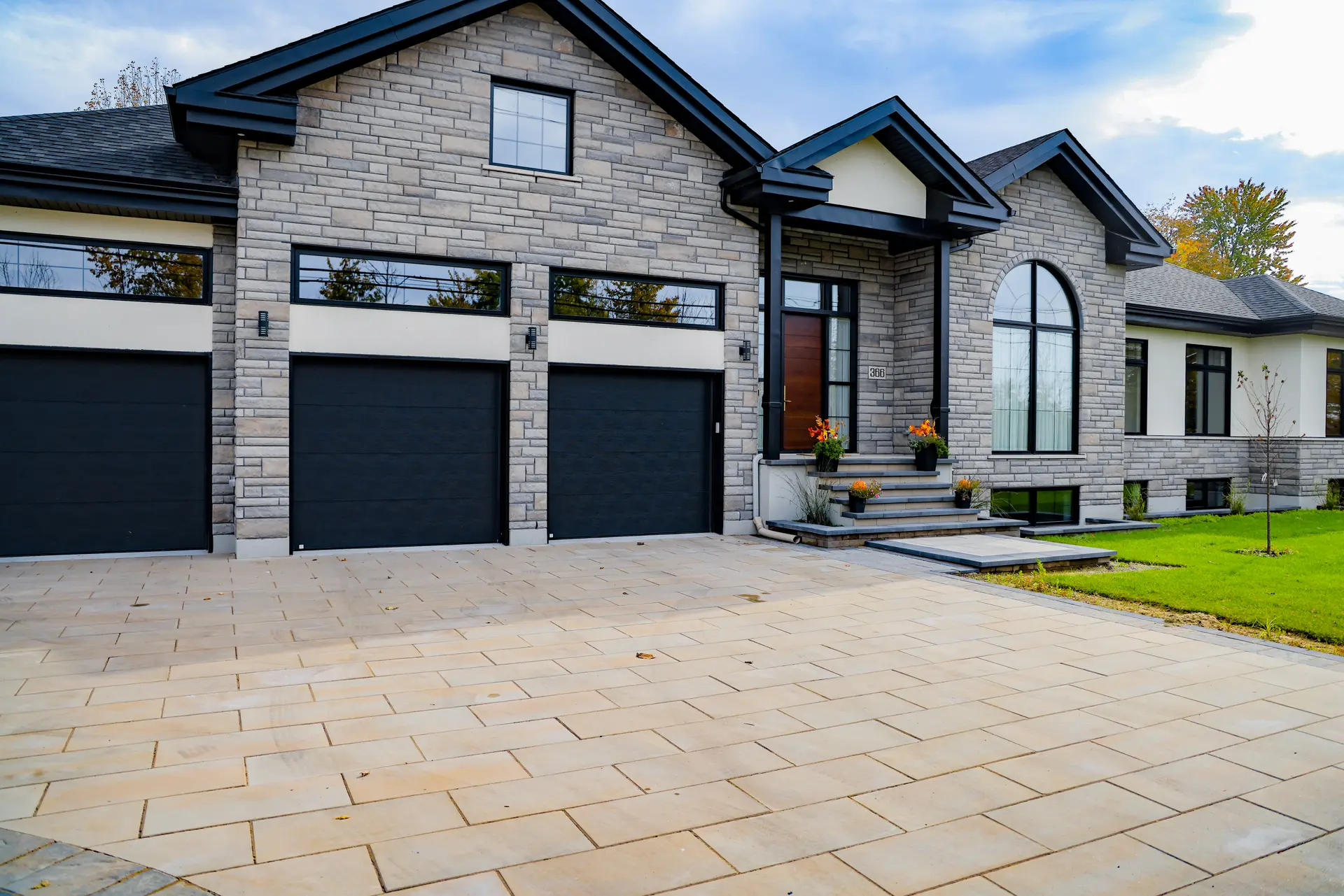 Modern house with stone facade, three black garage doors, and an entrance featuring large beige interlocking pavers, surrounded by a green lawn.