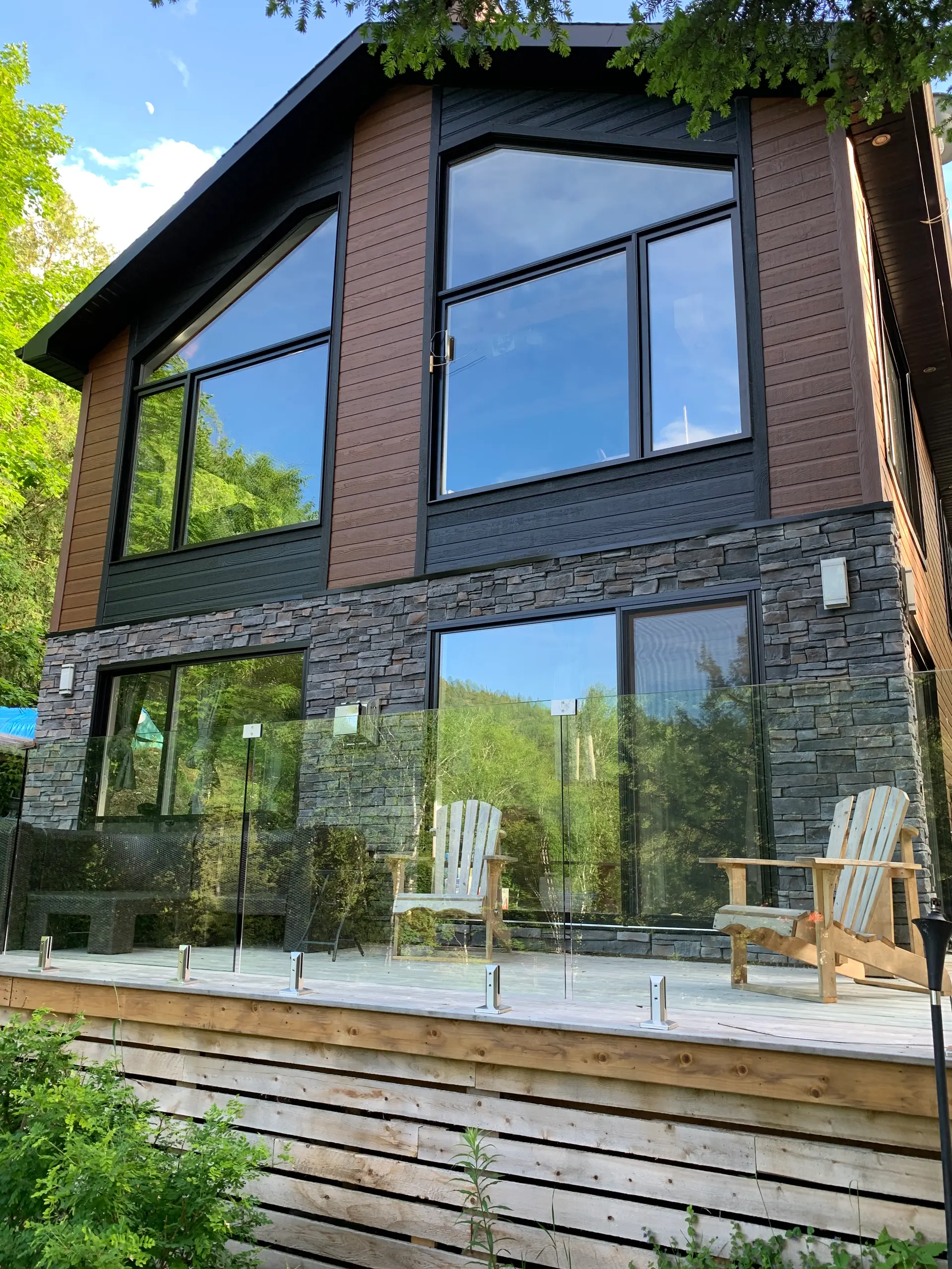 Modern house with wood siding, natural stone walls, large panoramic windows, and a wooden deck with a glass railing.