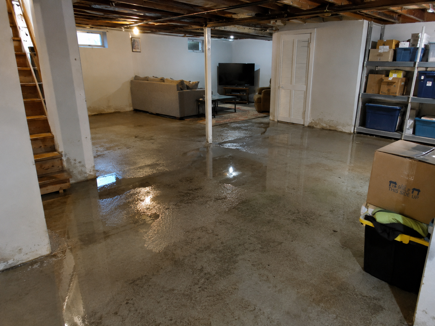 Flooded basement with standing water on concrete floor, damp walls, wooden stairs and residential storage area.