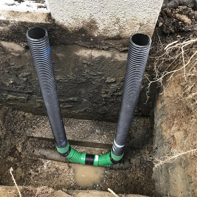 Installation de drains verticaux et de raccords de drainage au pied d’une fondation excavée avec sol humide, eau stagnante et mur de béton apparent