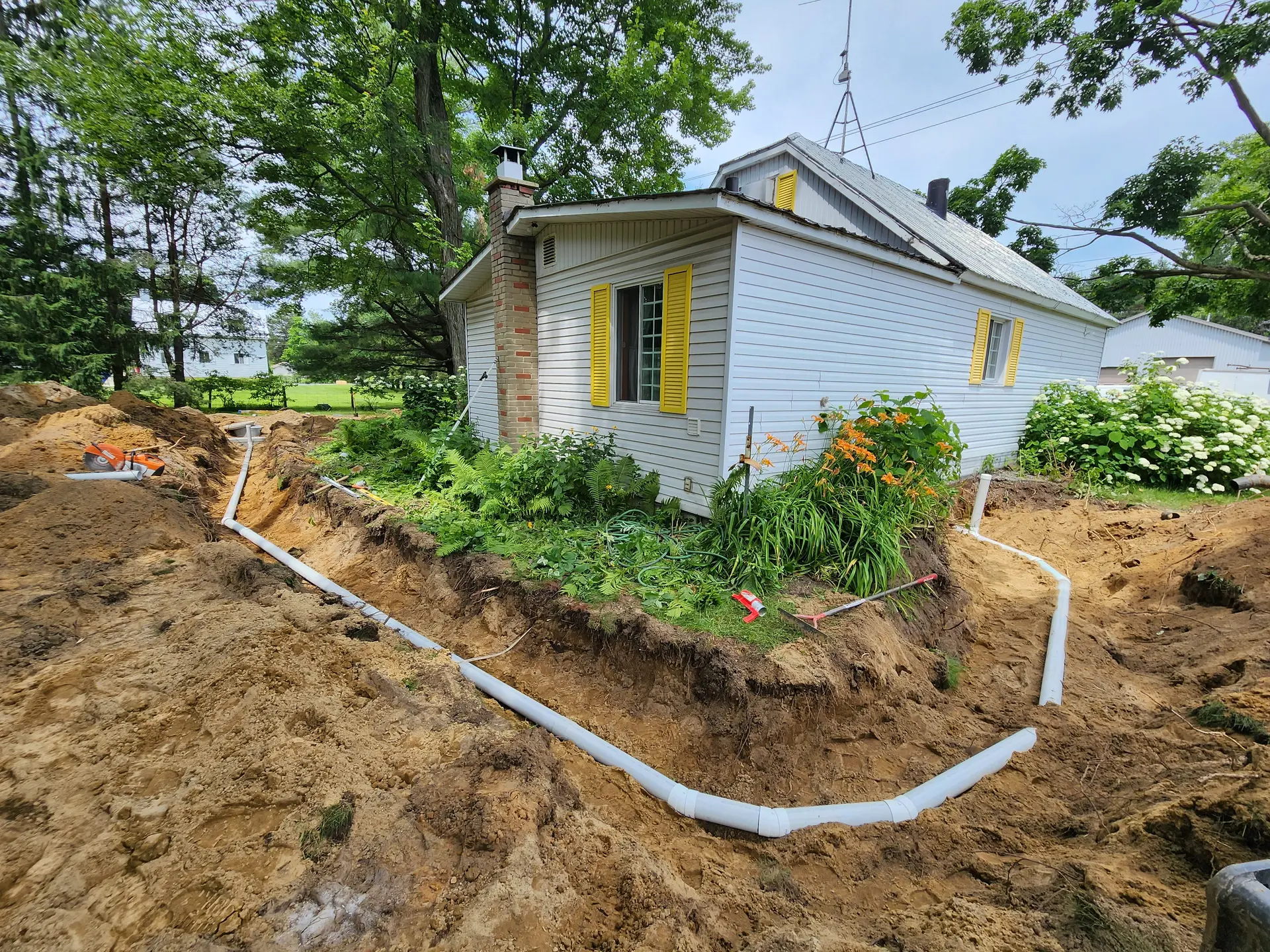 Installation de drainage extérieur avec tuyaux blancs autour d’une maison résidentielle excavée, jardin fleuri et aménagement paysager existant