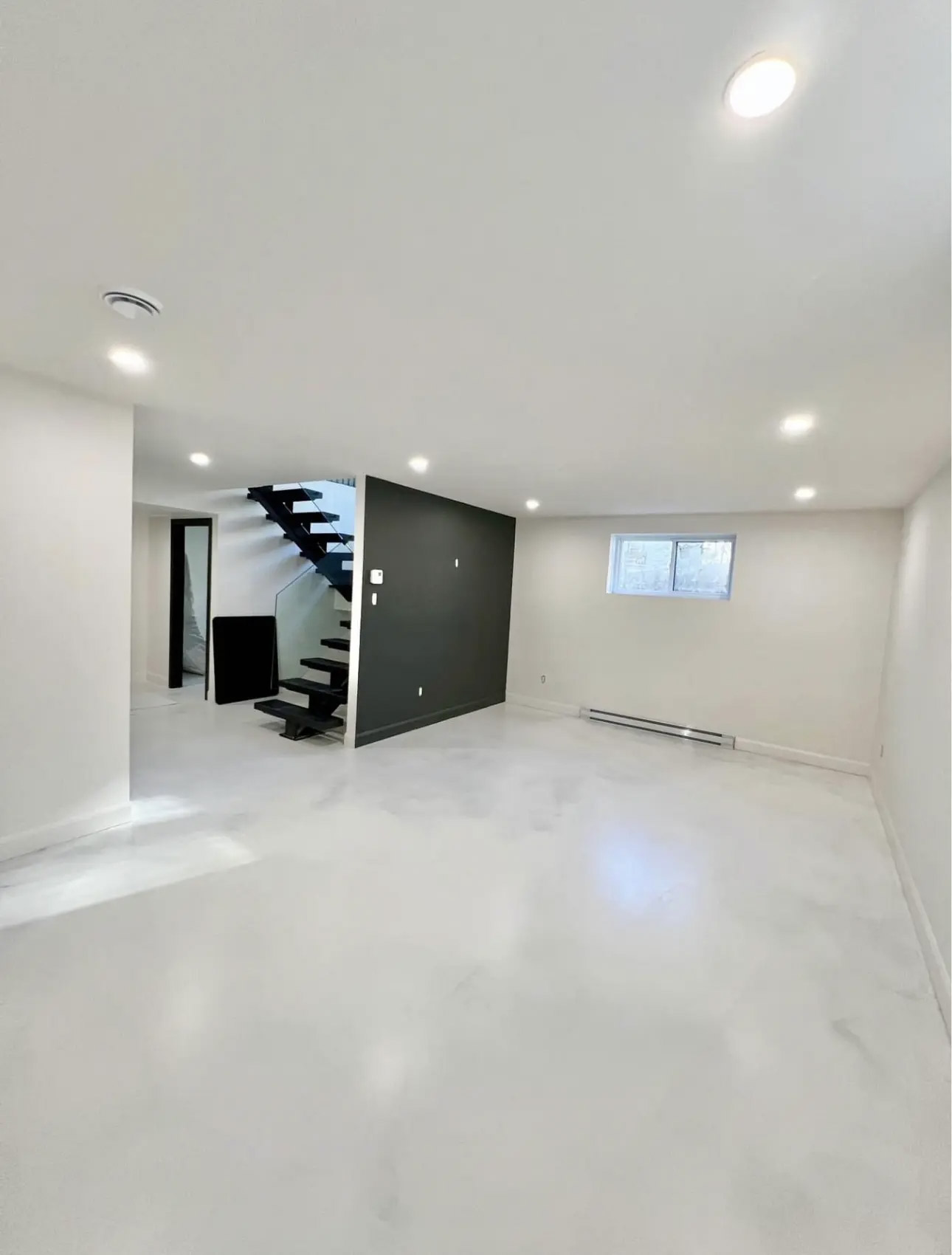 Bright renovated basement with glossy white epoxy flooring, modern black staircase, dark accent wall, recessed lights and low window.