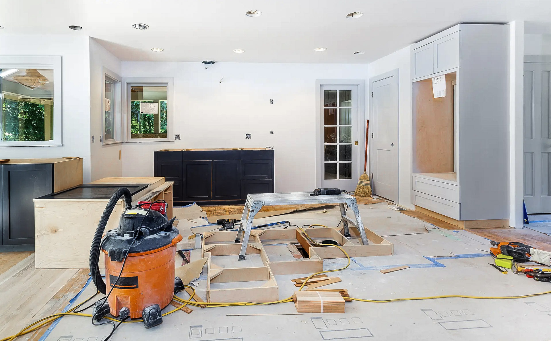 Cuisine intérieure en cours de rénovation avec armoires partiellement installées, outils de chantier, aspirateur, escabeau, protections au sol et éléments de menuiserie, illustrant des travaux de rénovation résidentielle, d’installation de cuisine et de finition intérieure