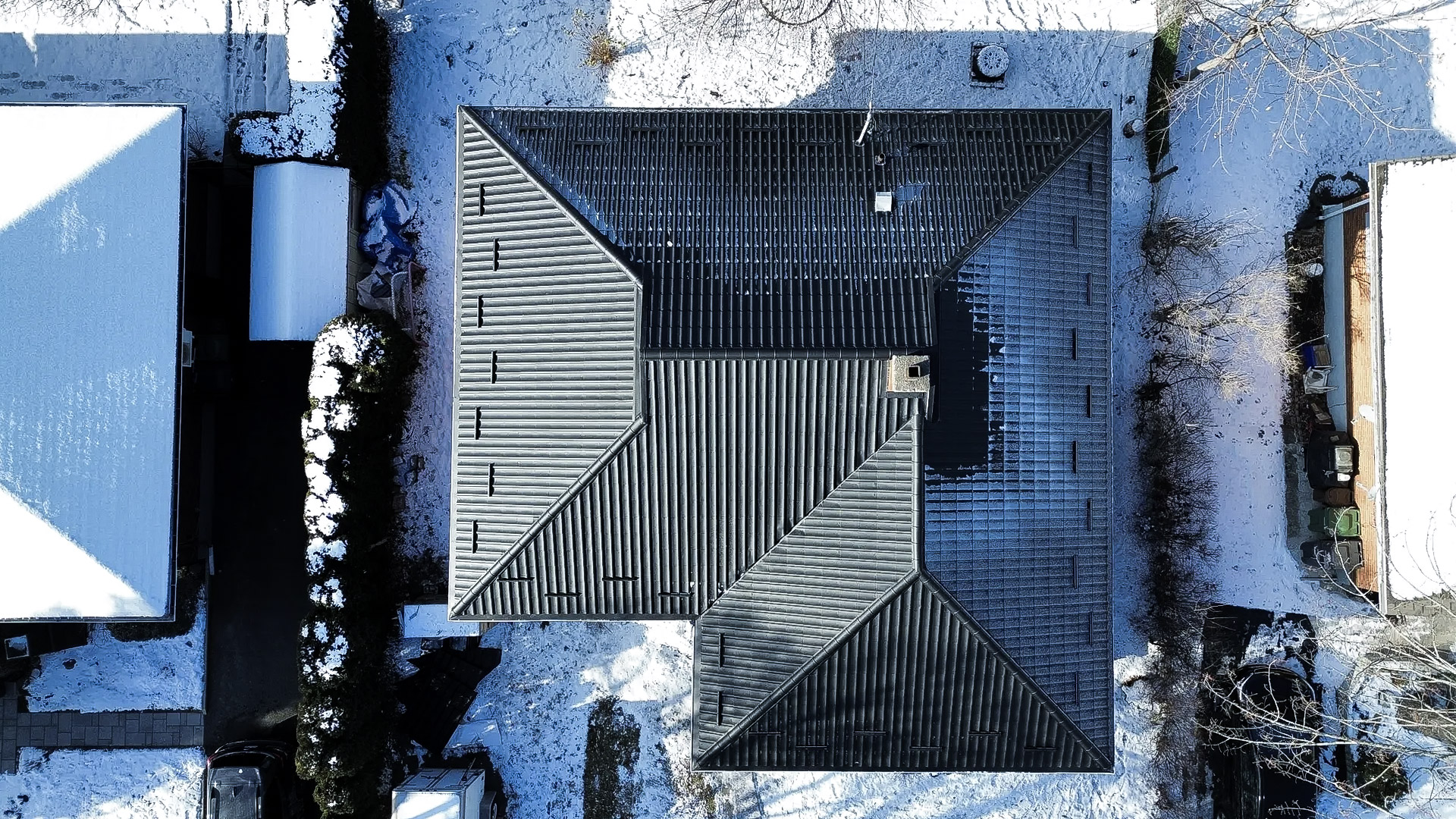 Vue aérienne d'une toiture en tôle noire sur une maison résidentielle entourée de neige en hiver, avec des arbres nus et des allées enneigées.