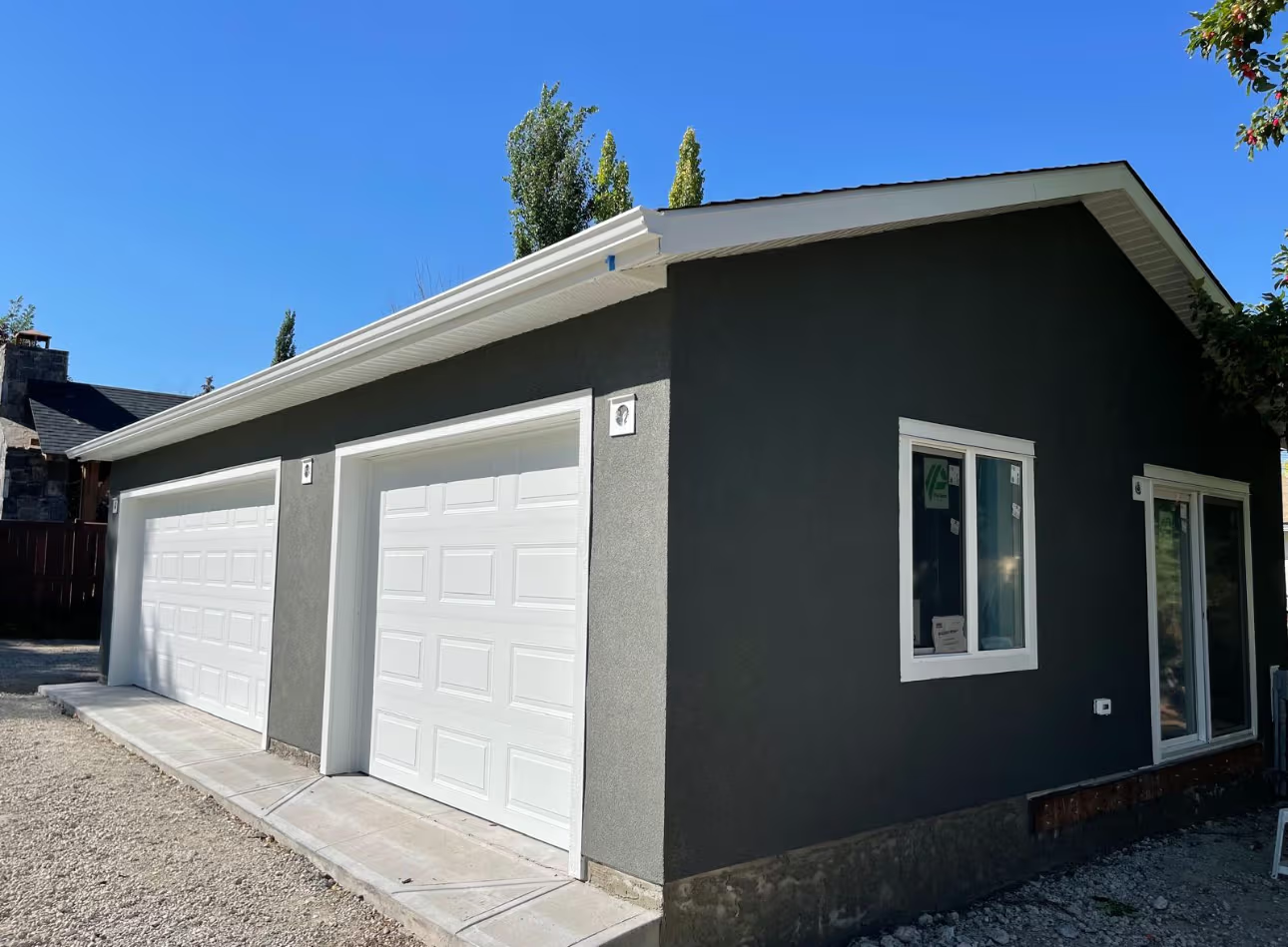 Garage résidentiel gris foncé avec deux portes de garage blanches, soffites et fascias blancs, fenêtres récentes, revêtement extérieur en crépi et entrée en gravier